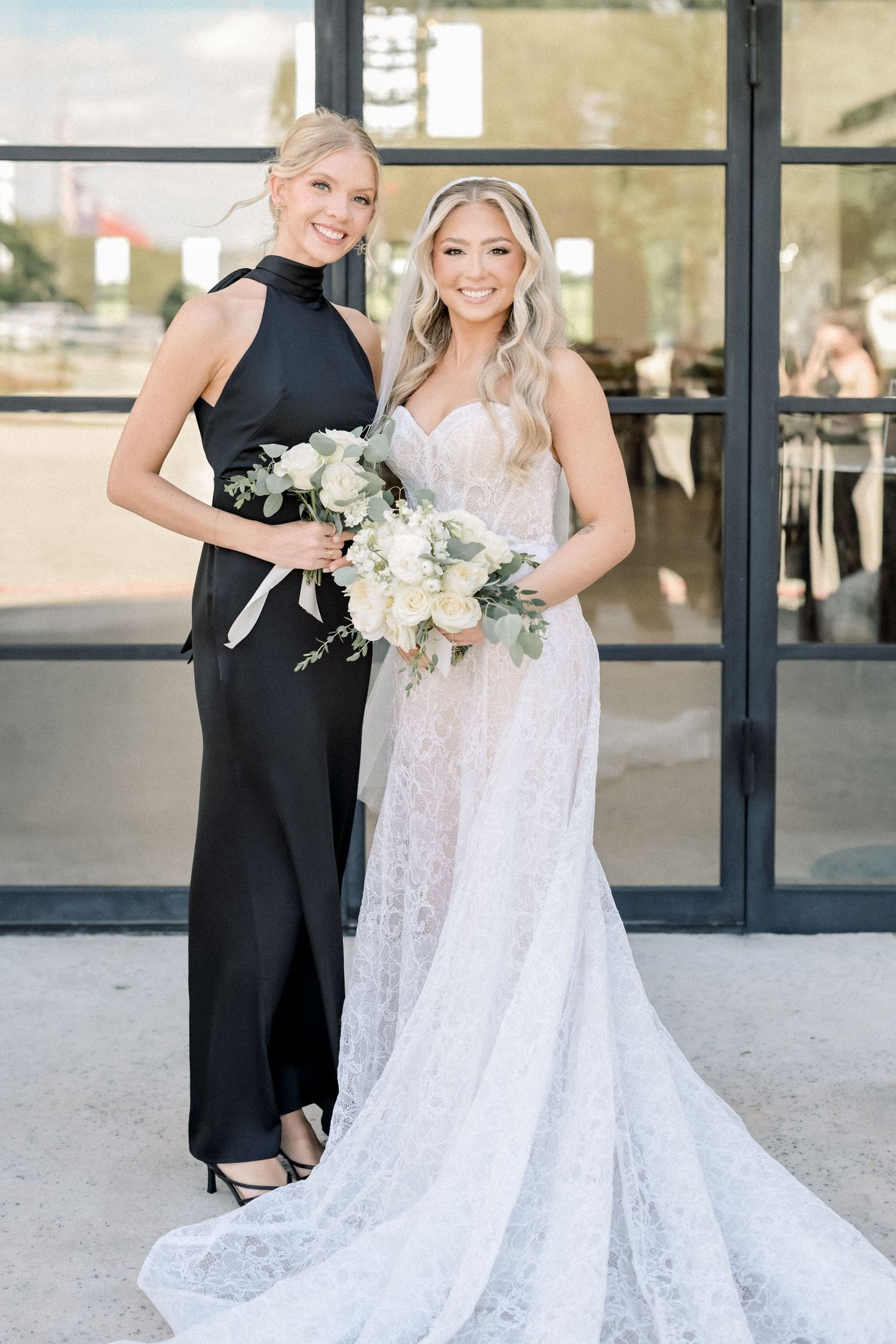 Bride in lace gown with long train poses with bridesmaid in black halter jumpsuit outdoors.
