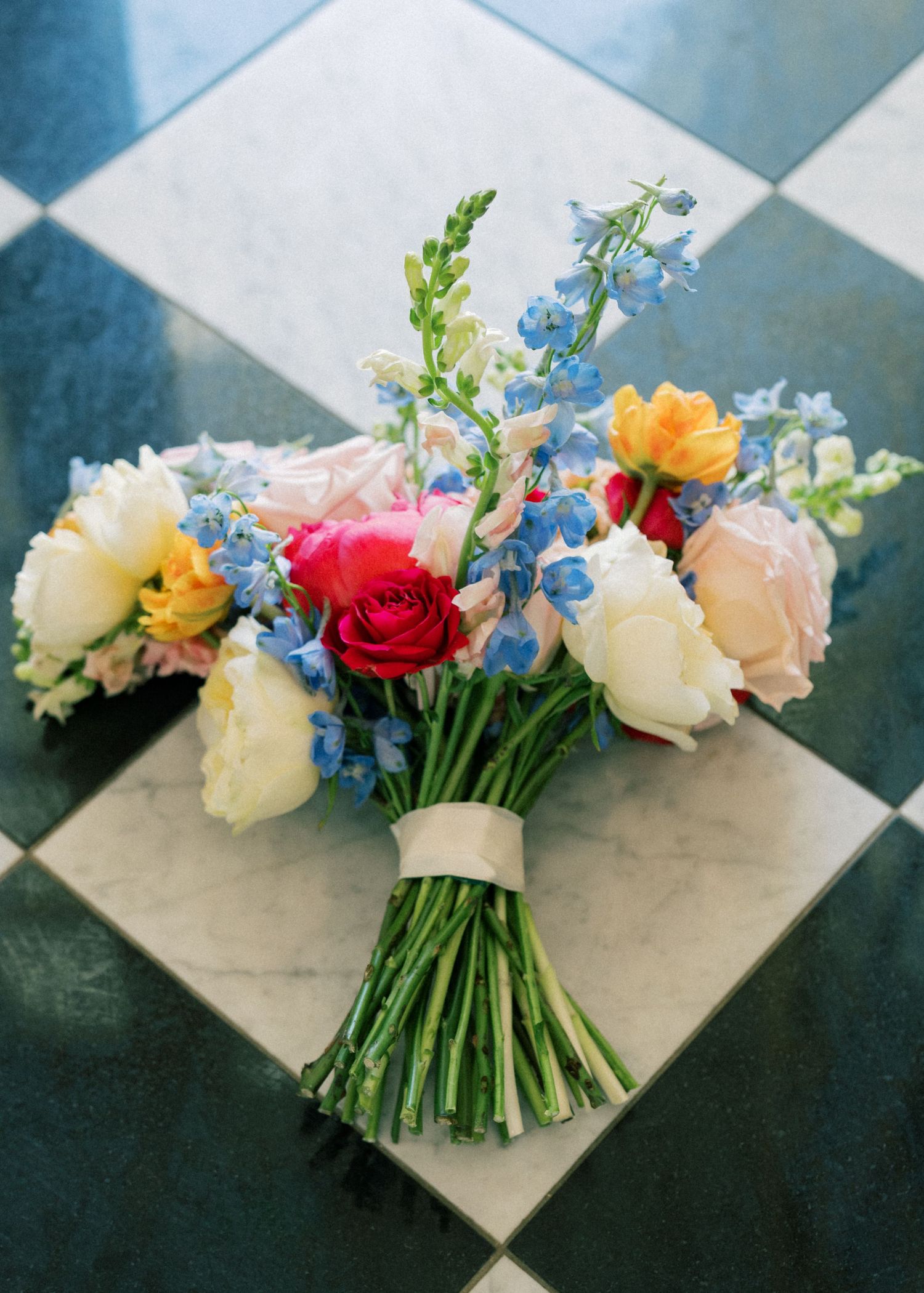Colorful bridal bouquet with pink roses, blue delphiniums, and white peonies resting on a checkered marble floor.