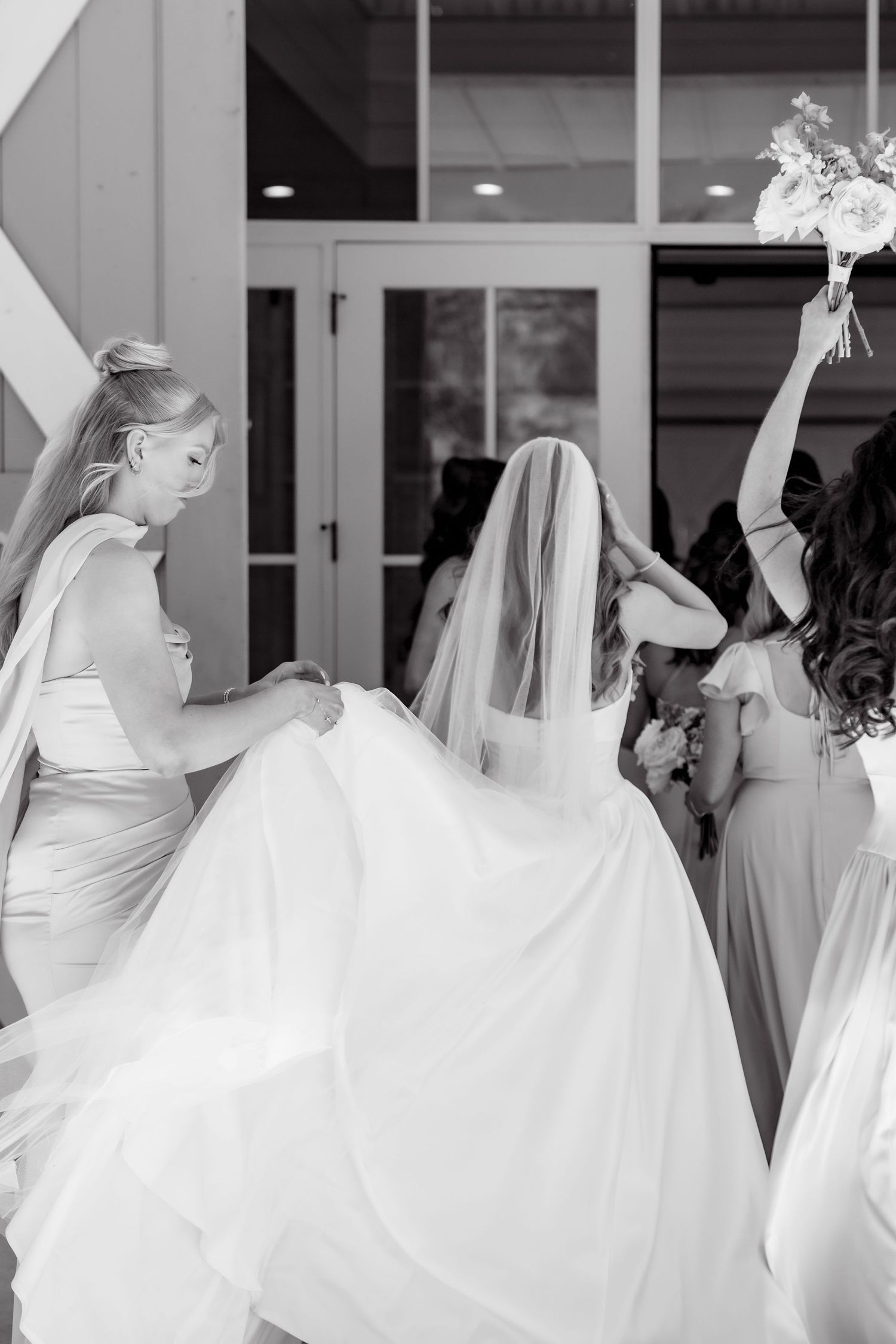 Black and white photo of bridesmaids helping bride with veil and train outside wedding venue.