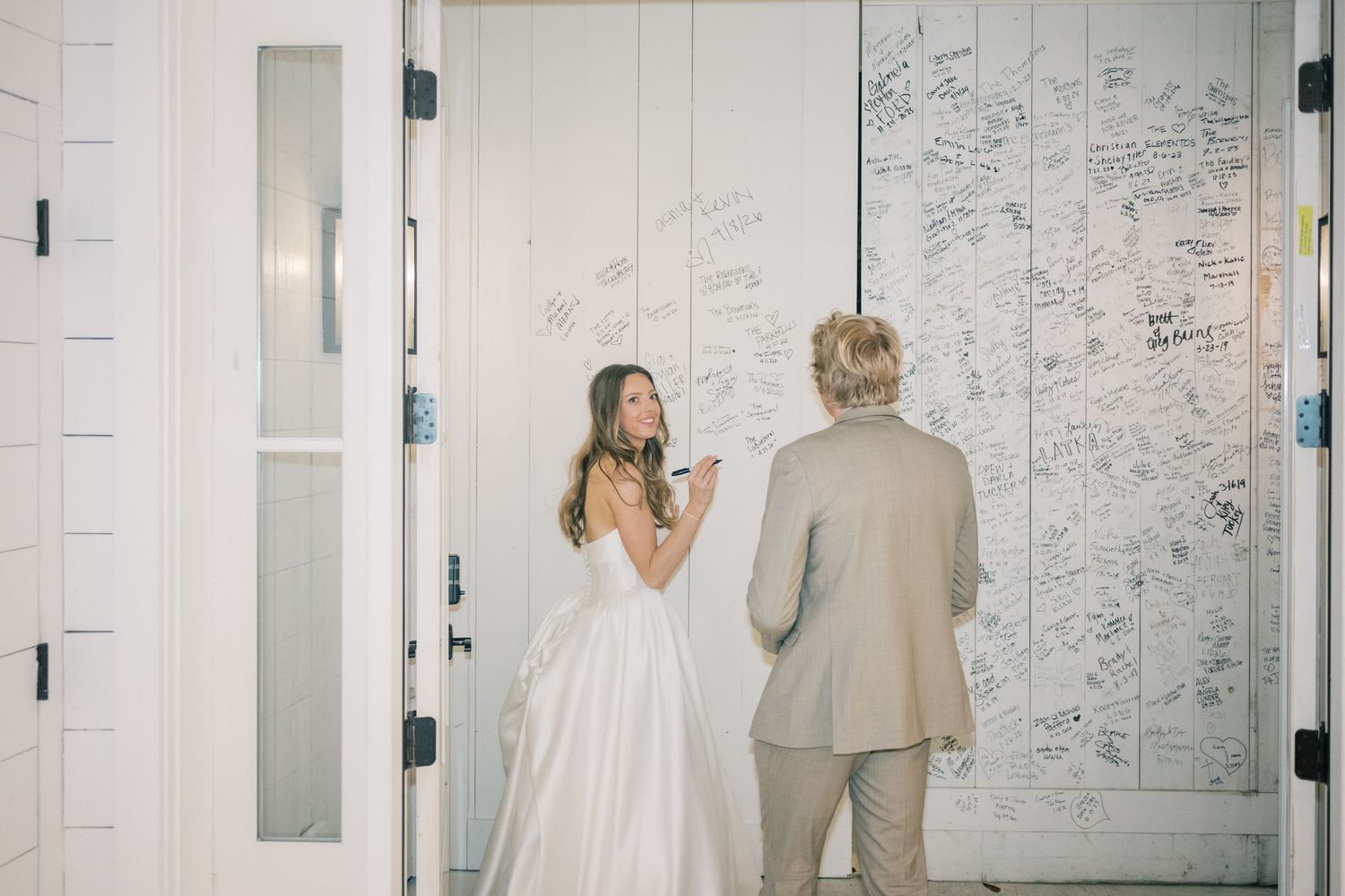 Bride and groom signing a large white wall covered in wedding guest messages at their reception.