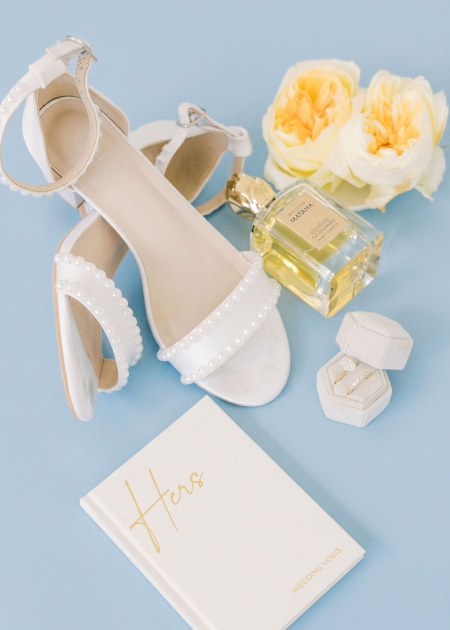 White bridal heels, engagement ring, perfume bottle, and yellow roses arranged on a light blue background.