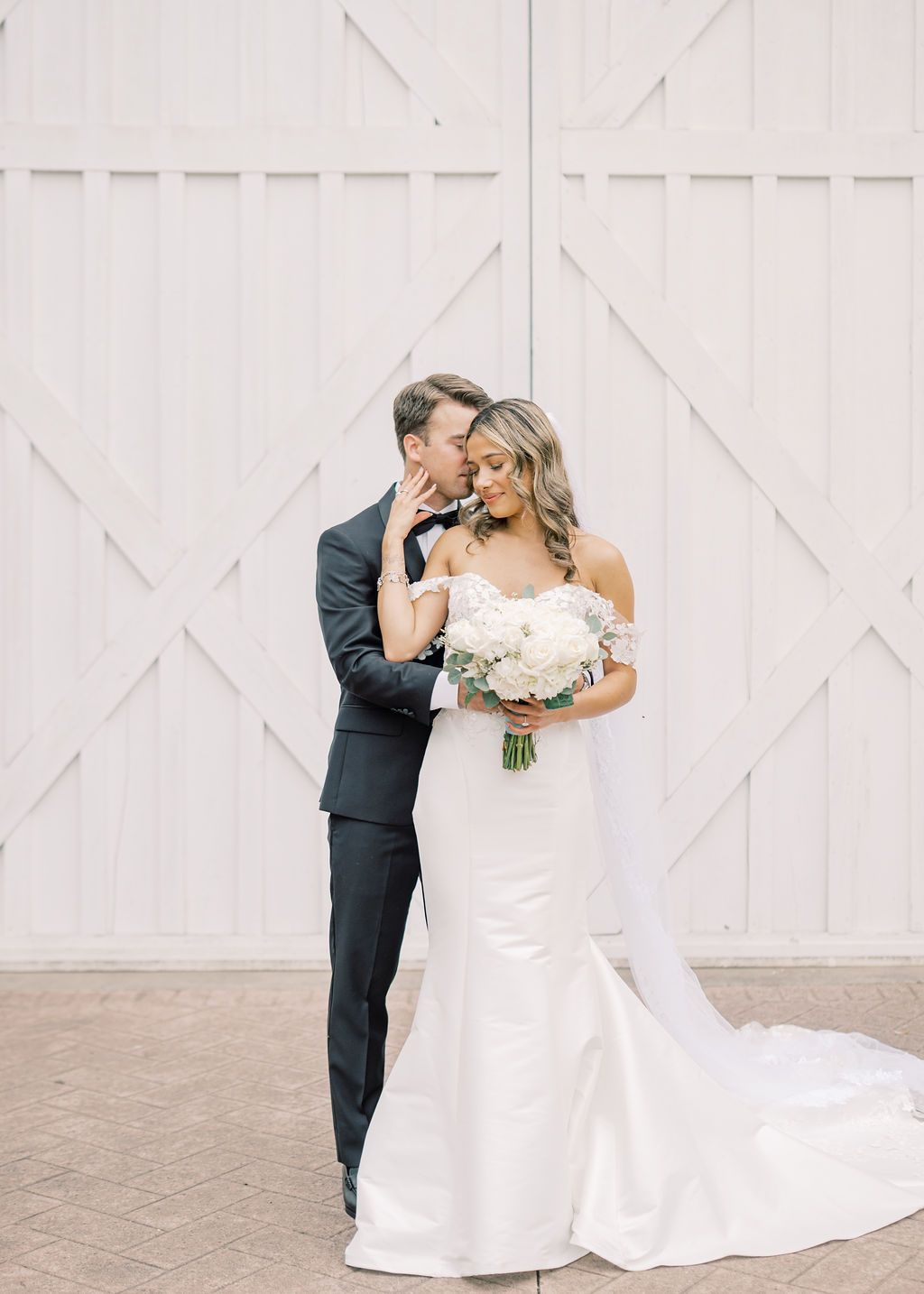 Groom kisses bride holding white bouquet in mermaid gown outside white barn doors.