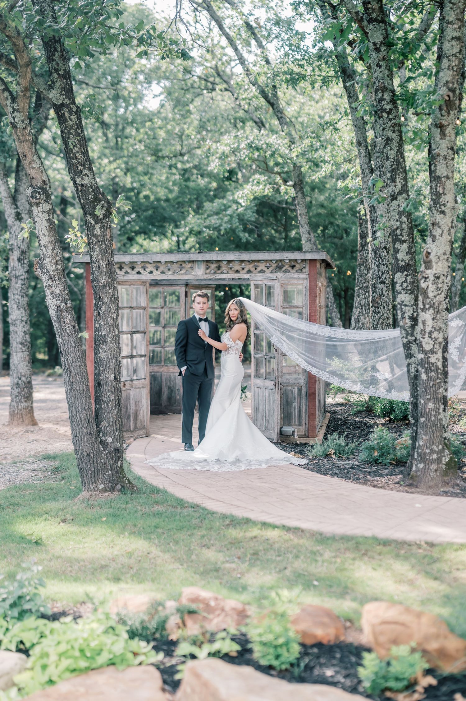 Bride and groom standing together in a lush garden setting with veil flowing in the breeze.