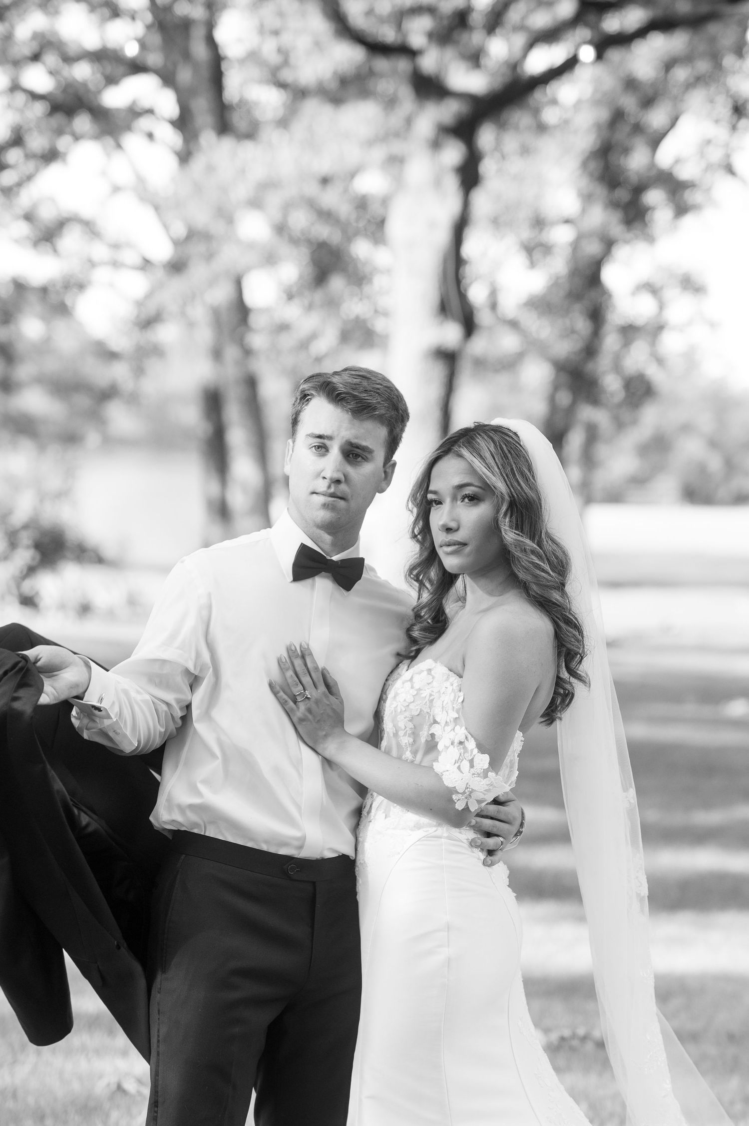 Black and white portrait of bride and groom posing outdoors under trees.