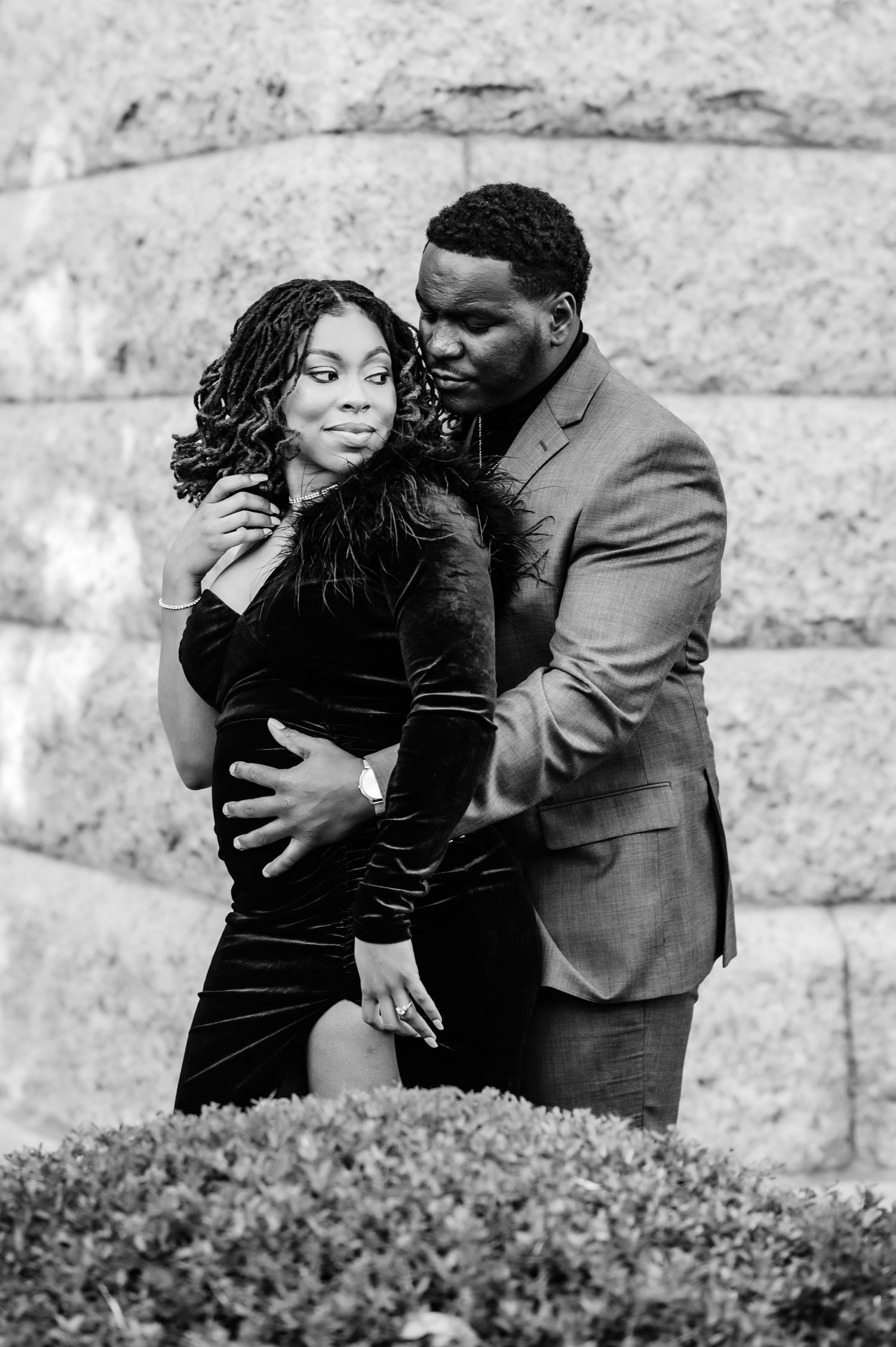 Black and white photo of a couple embracing romantically in front of a stone wall, man in suit kissing womans cheek.