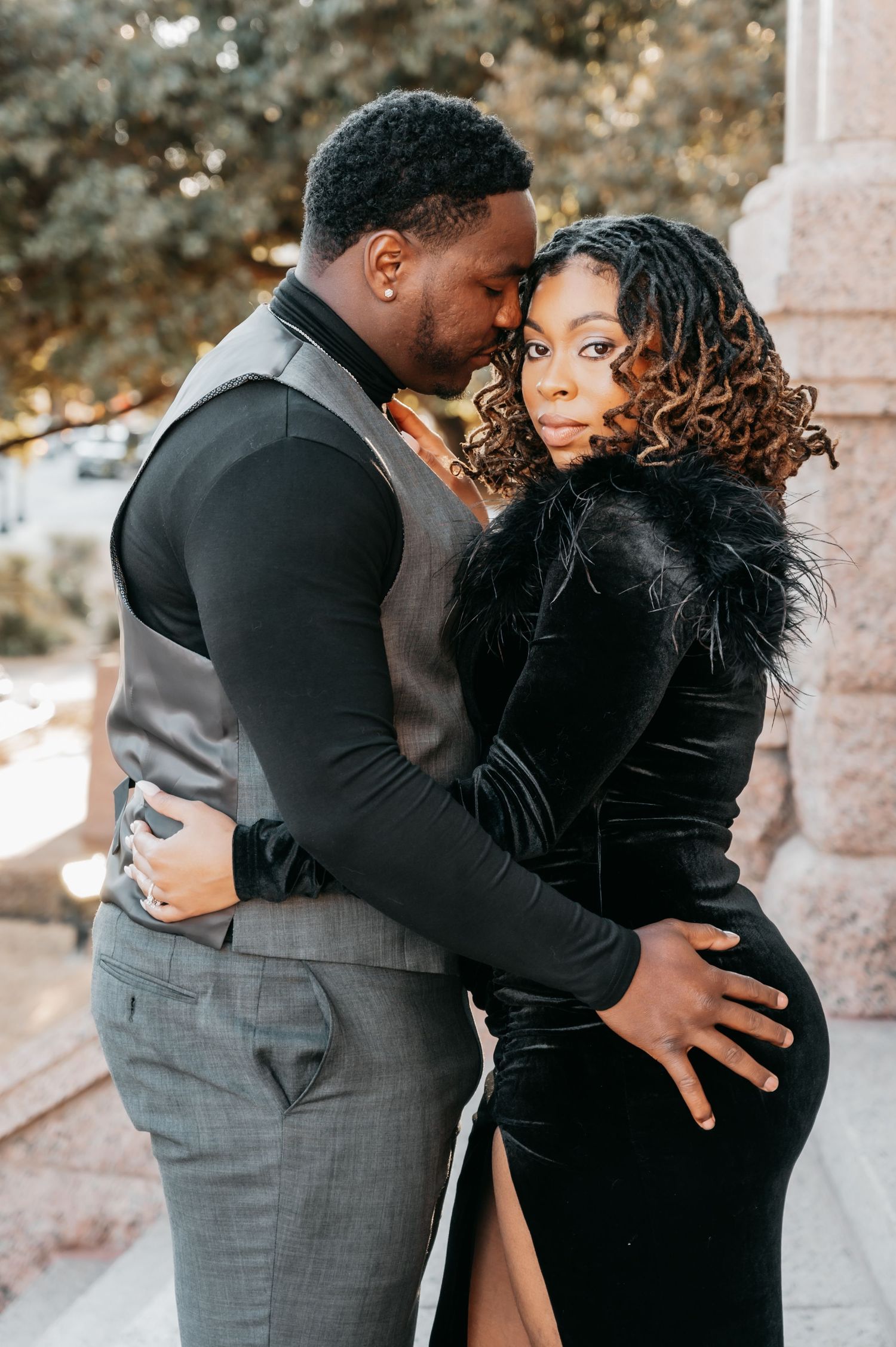 A couple in elegant black and gray formal attire share an intimate embrace outdoors during an engagement photo session.
