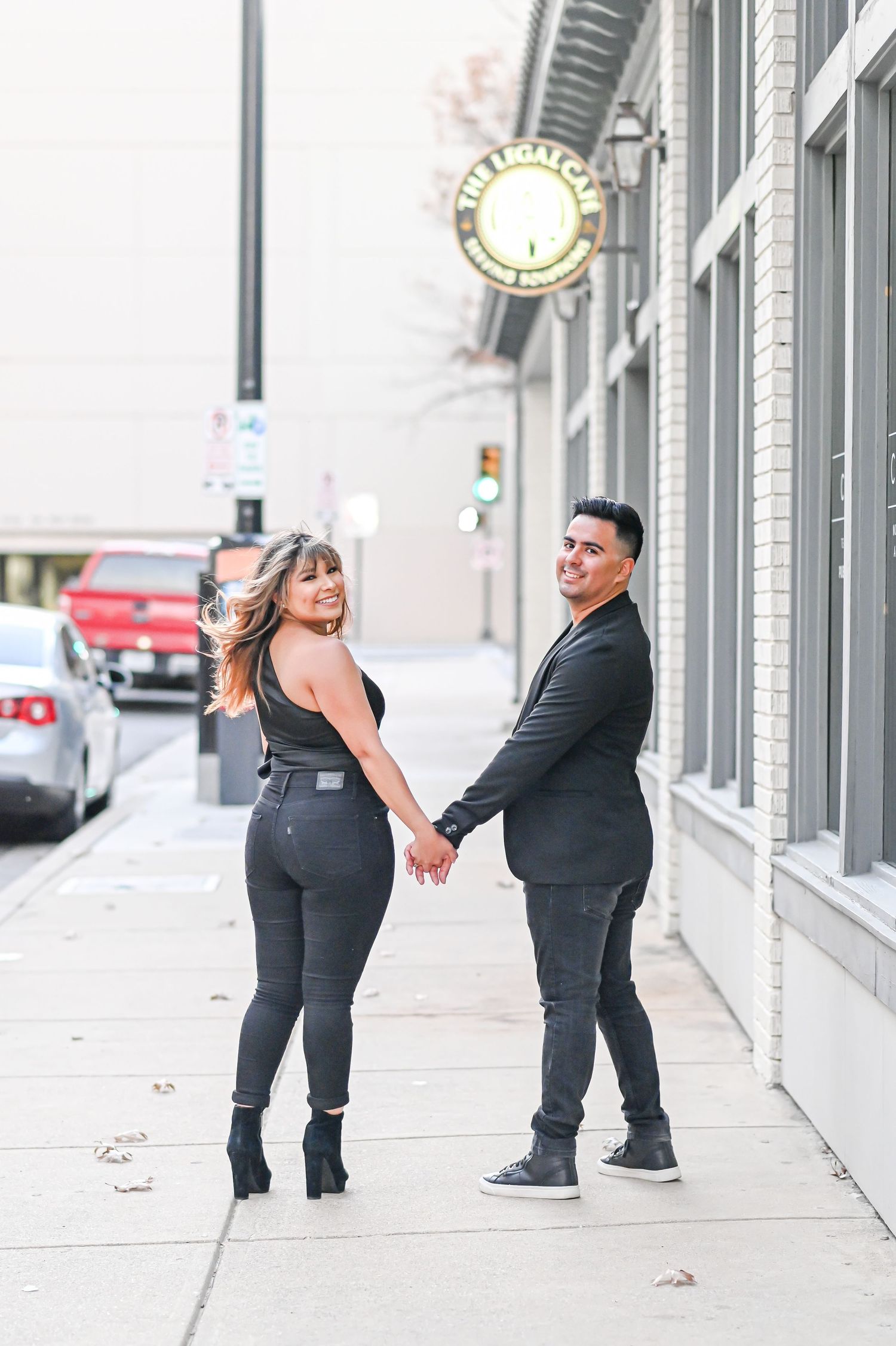 A stylish couple holds hands on a city sidewalk, posing and looking back at the camera.