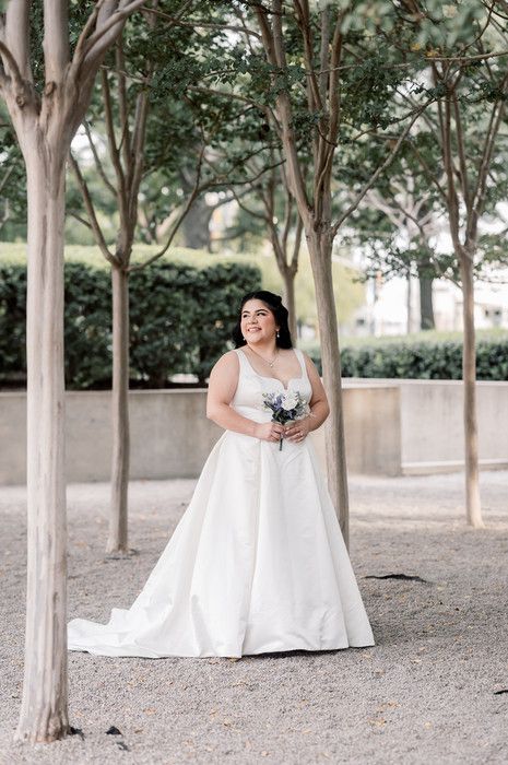 Bride in a white A-line gown holding a bouquet, smiling among trees in an outdoor garden setting.