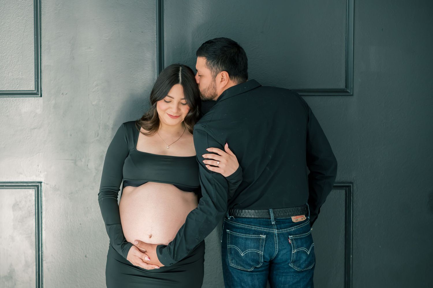 Couple sharing a tender maternity photo moment, man kissing pregnant woman's head against a dark gray paneled wall.