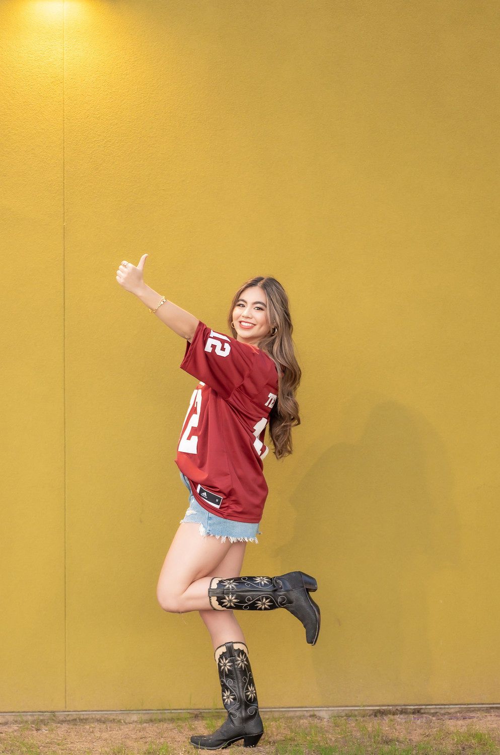 Woman in Texas jersey, denim shorts, cowboy boots, giving thumbs up against yellow wall.