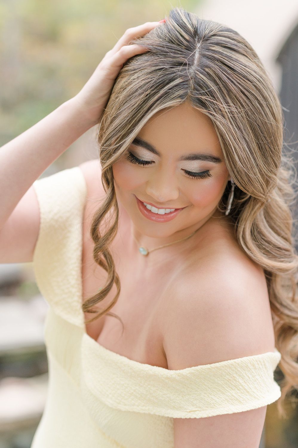 Young woman in off-shoulder yellow dress with wavy blonde hair, elegant makeup, and delicate necklace, smiling softly. Wichita Falls HS