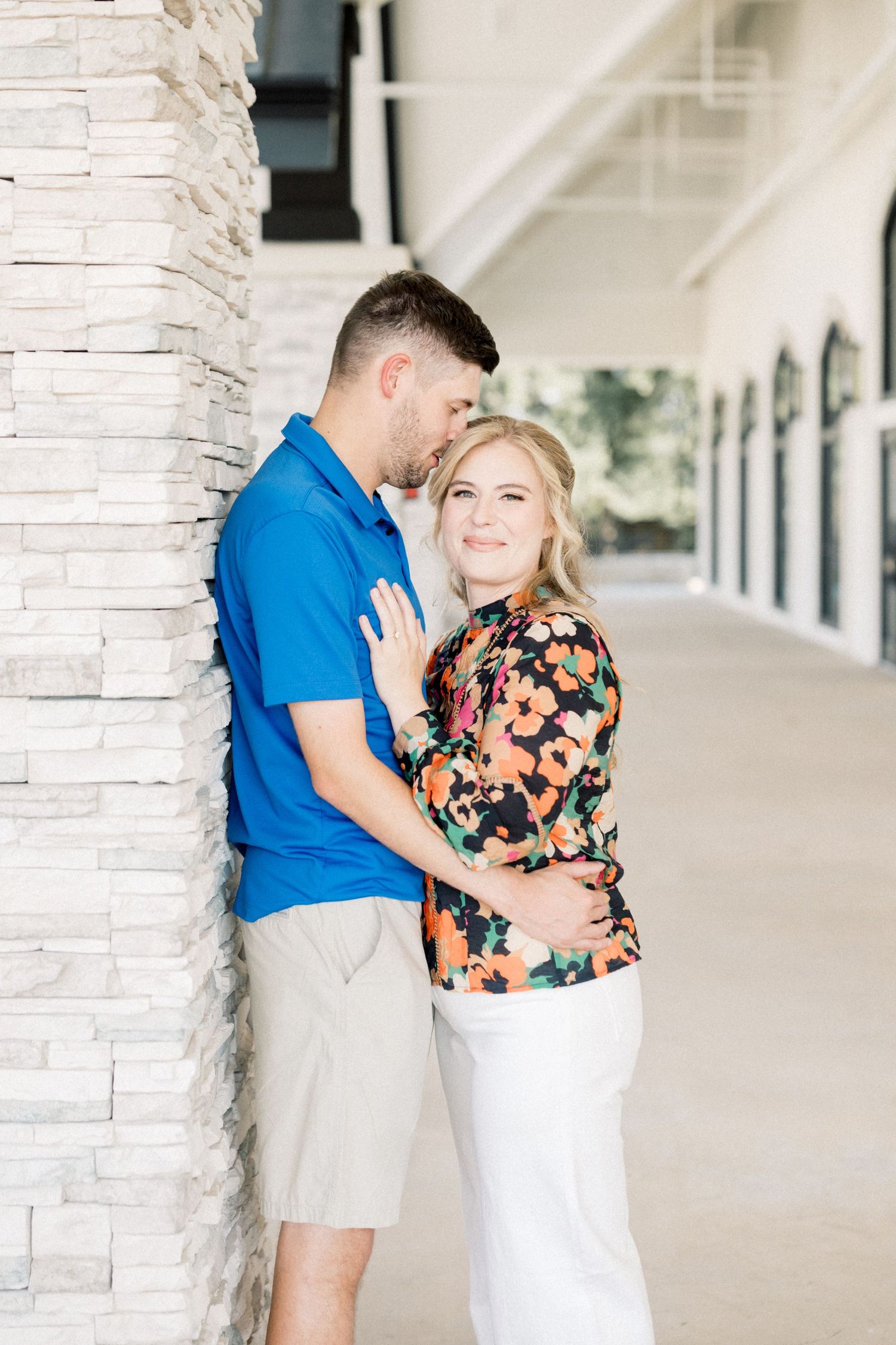 Couple embracing by stone pillar, man in blue polo kissing woman in floral top wearing white pants.