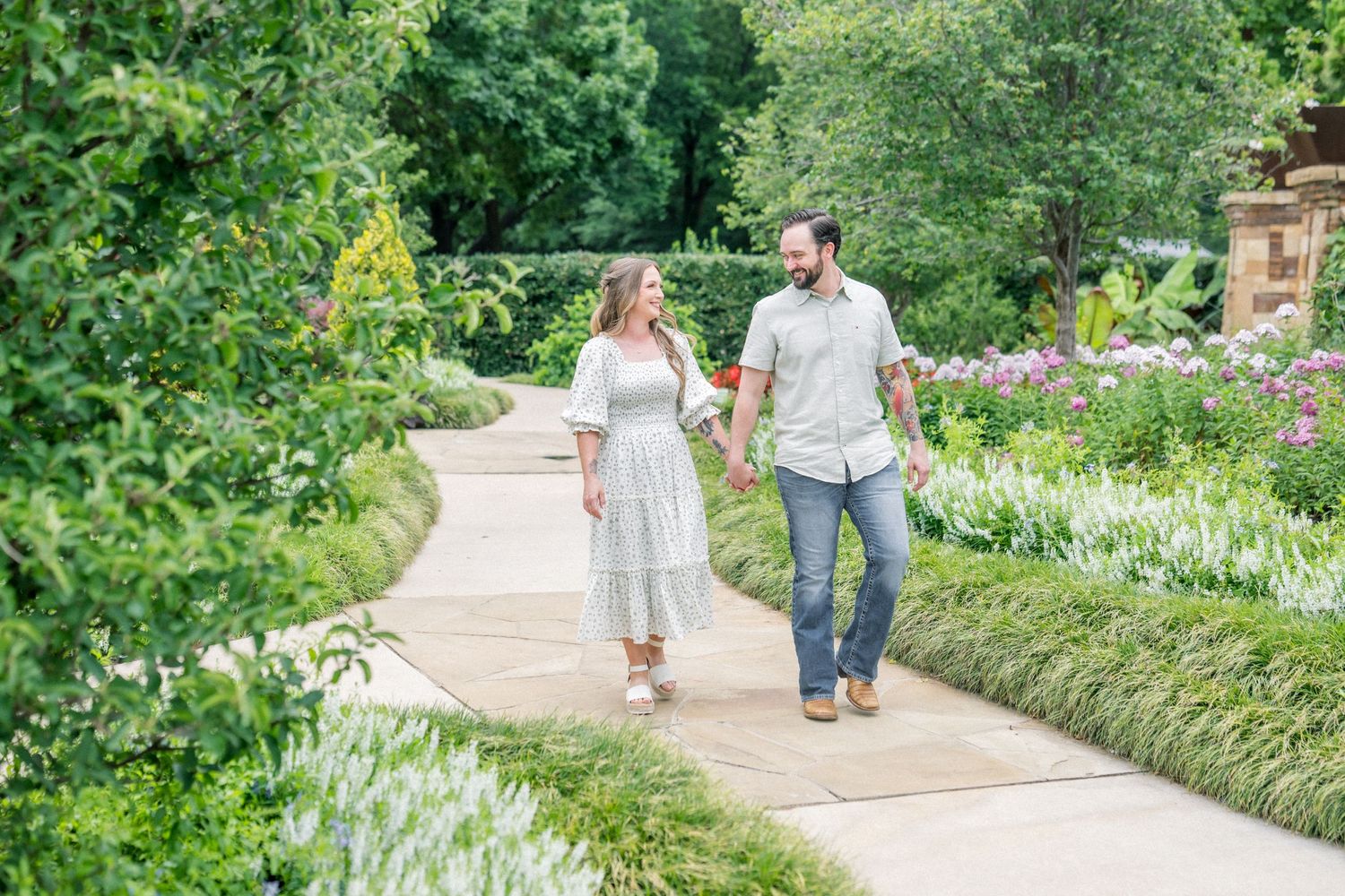 Couple holding hands and walking on a garden path surrounded by lush greenery and colorful flowers.
