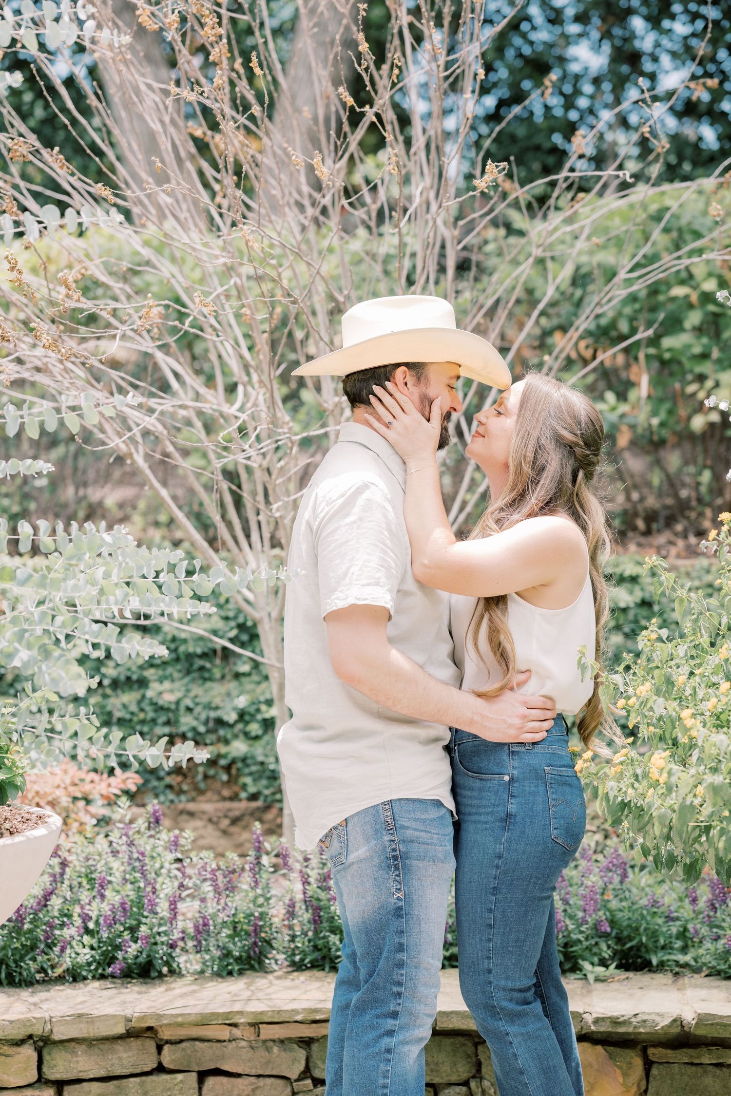 Couple sharing a kiss outdoors in a lush garden, man in cowboy hat and woman in white top and jeans.