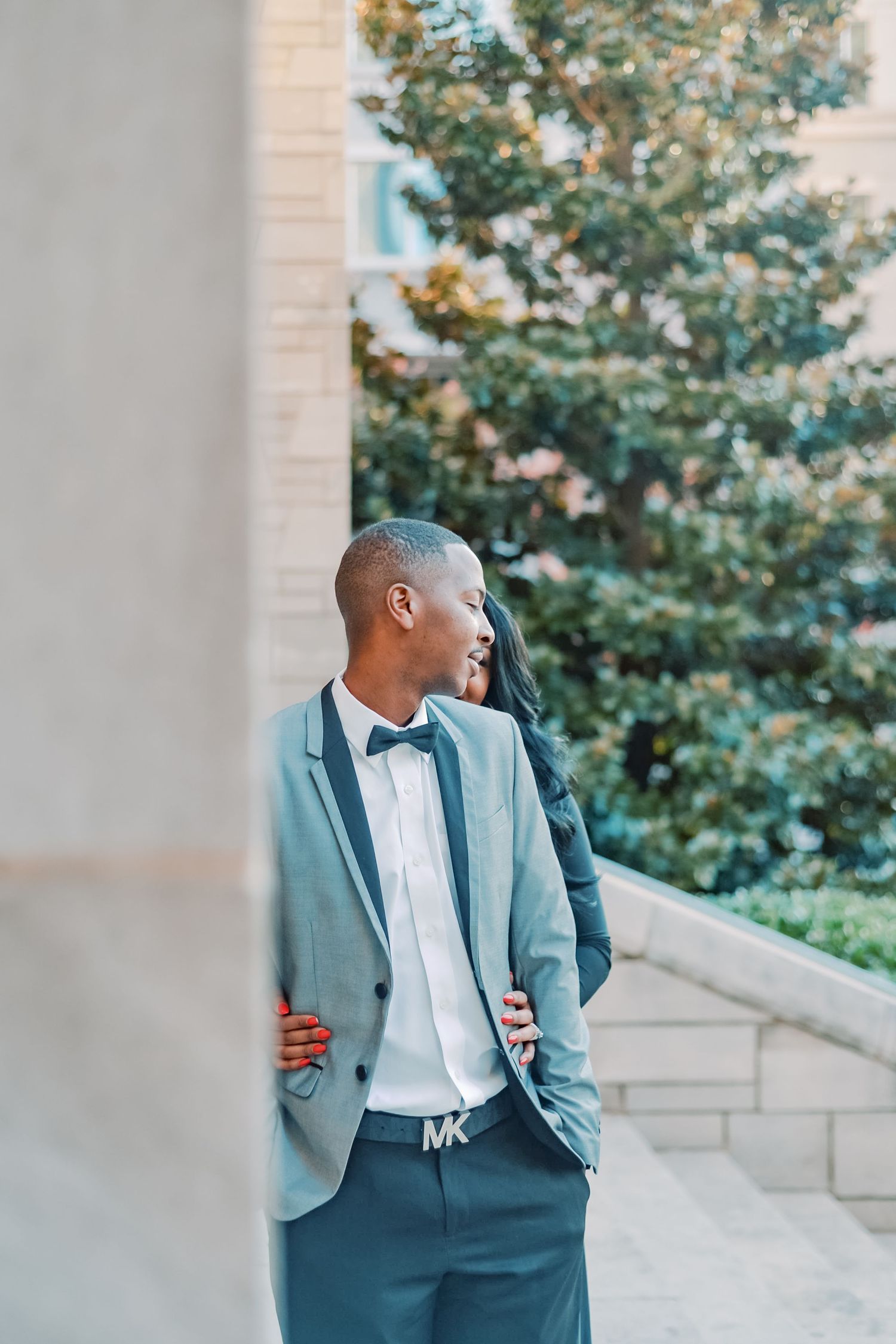 Stylish young Black man in light blue suit with bow tie and MK belt, smiling outdoors near stone column.