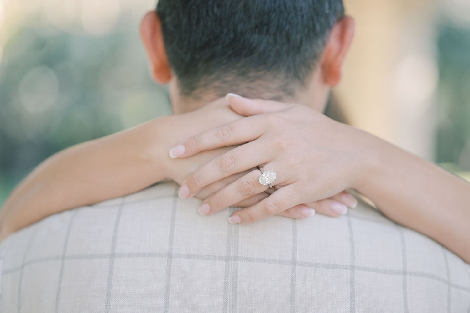 Woman's arms wrapped around a man's shoulders from behind, her hand showing a sparkling diamond engagement ring against a plaid shirt.