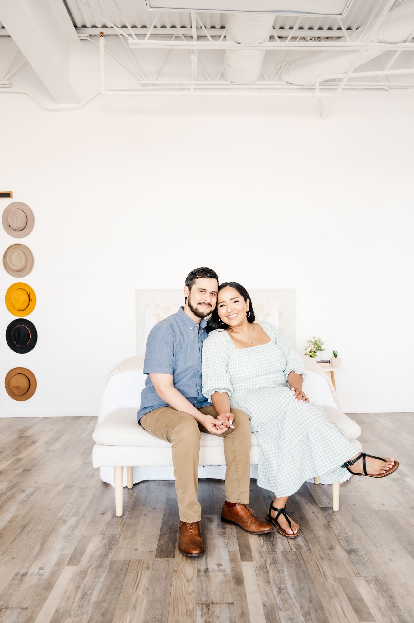 A romantic portrait in a bright studio with decorative hats on the wall and a white bench.