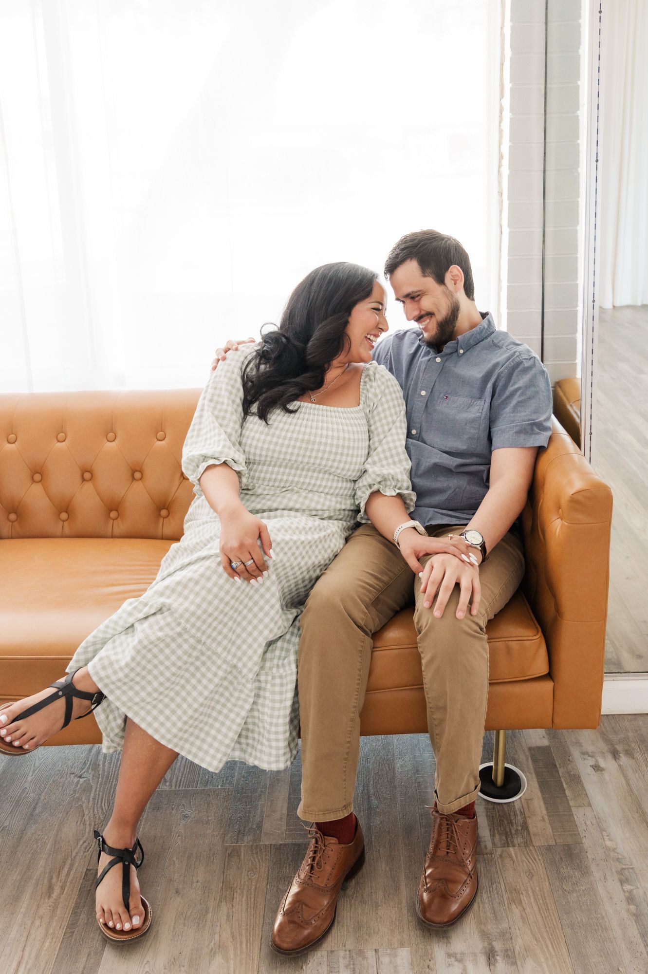 A couple laughs together while sitting on a camel-colored sofa near a bright window.
