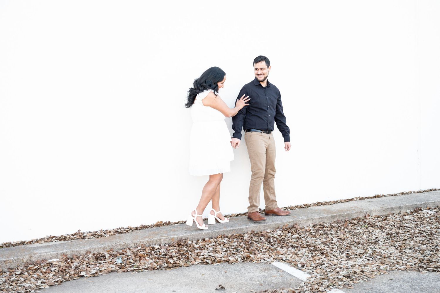 Couple in black and white attire shares candid moments along a white wall with fallen leaves.