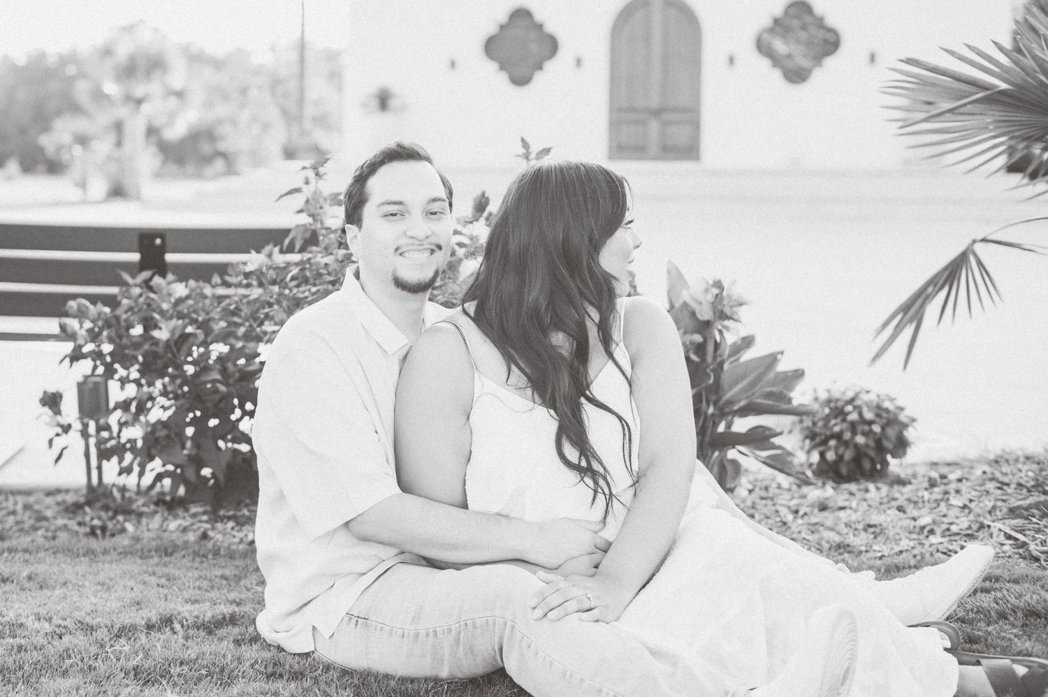 A couple in white outfits shares romantic moments during an outdoor photo session in a garden setting.