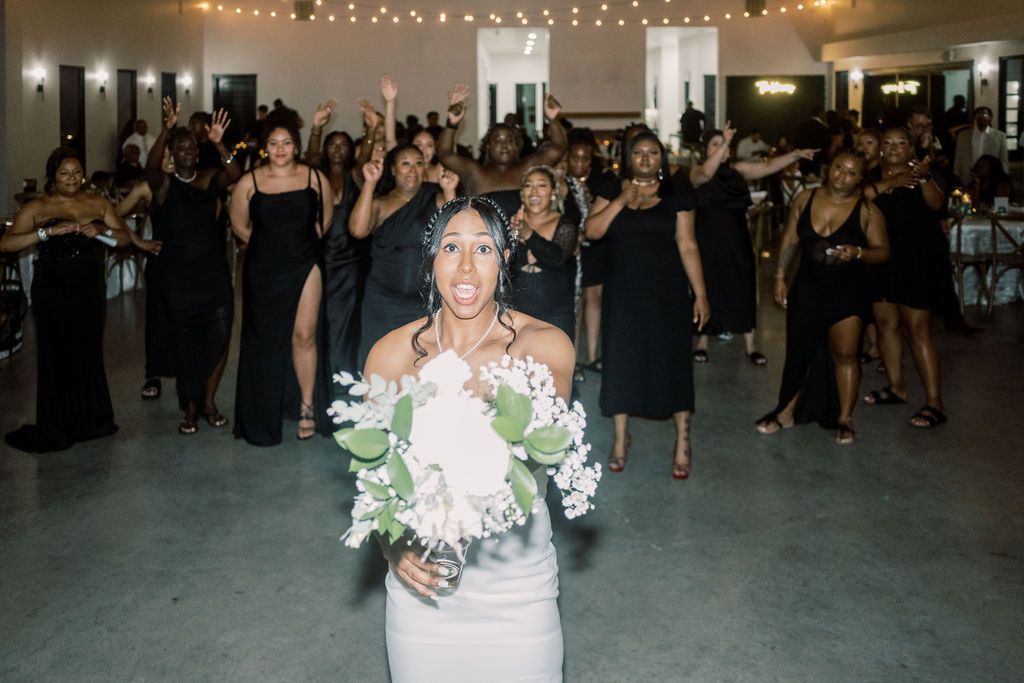 Bride in white gown holds bouquet while bridesmaids in black dresses celebrate behind her at wedding reception.