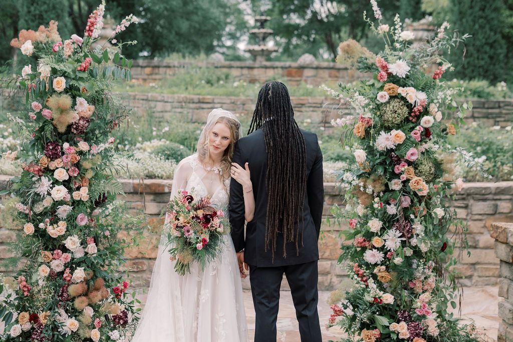 Bride and groom stand at floral arch with pink and peach blooms during outdoor wedding ceremony.