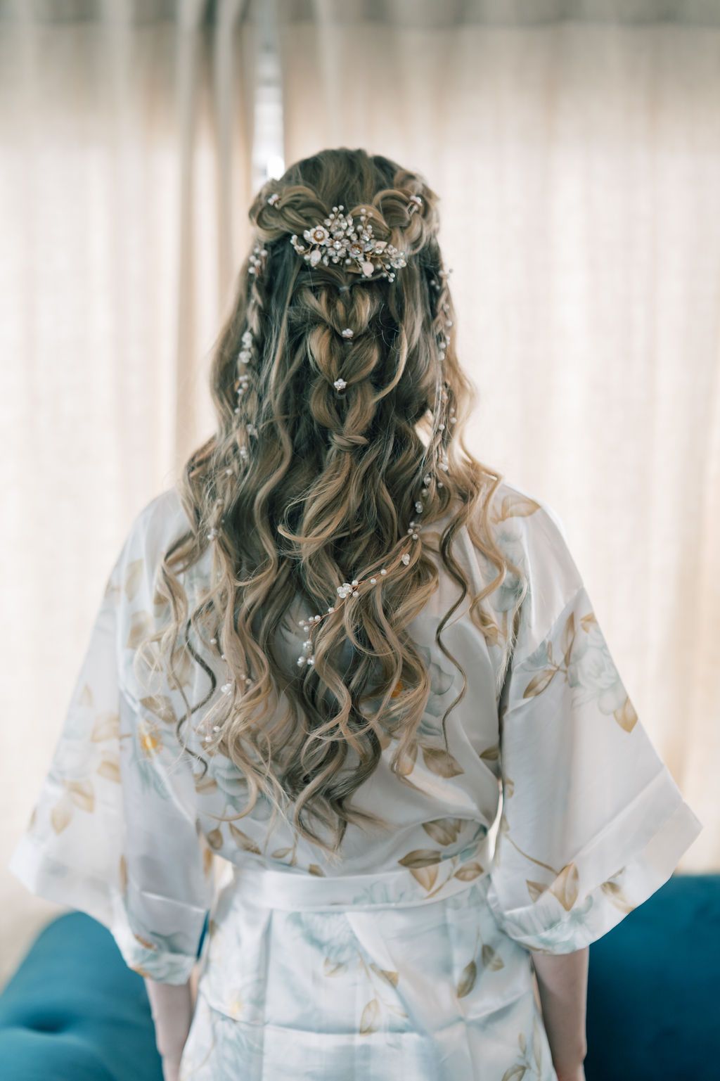 Bride with long curly hair in half-up style adorned with crystal hair vine, wearing floral robe before wedding.
