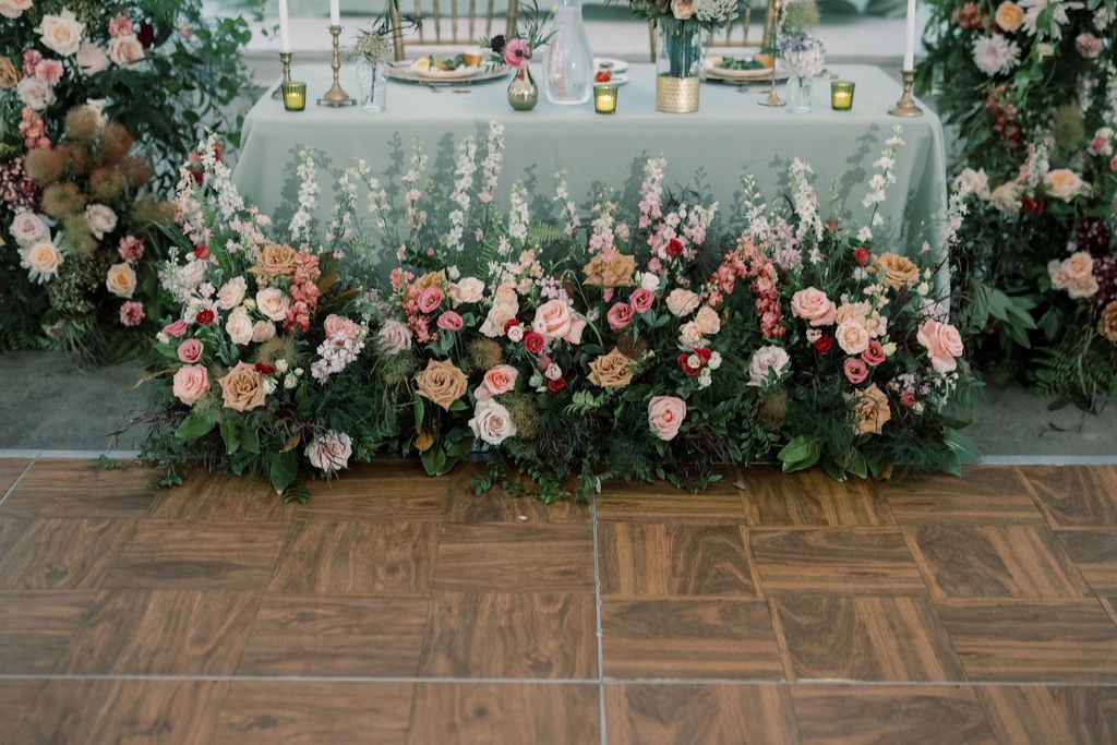 Elegant wedding head table adorned with lush pink and white floral arrangements and greenery against a sage green tablecloth.