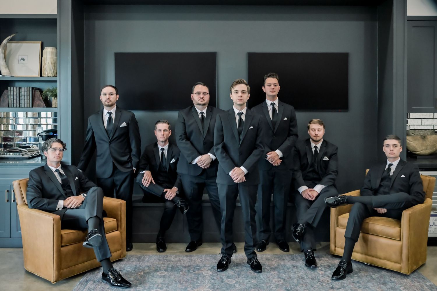 Eight groomsmen in matching dark suits pose formally in a modern lounge with leather chairs for a wedding party photo.