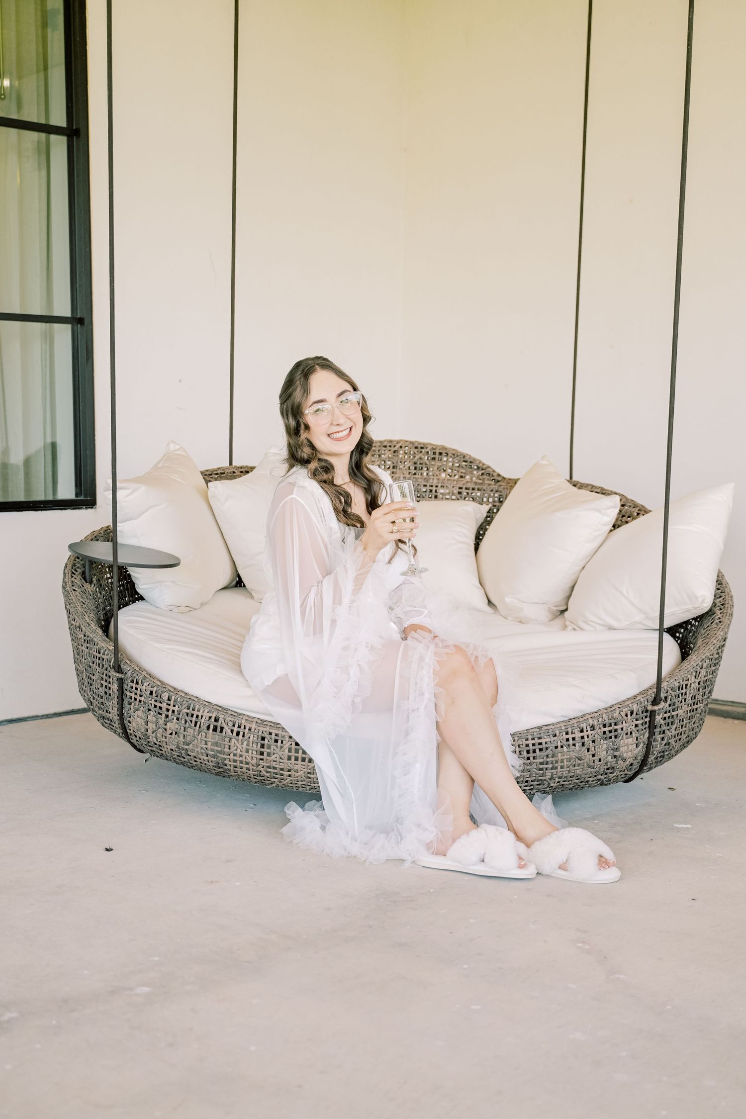 Smiling bride in white lace robe and fluffy slippers relaxing on a wicker swing chair holding a drink.