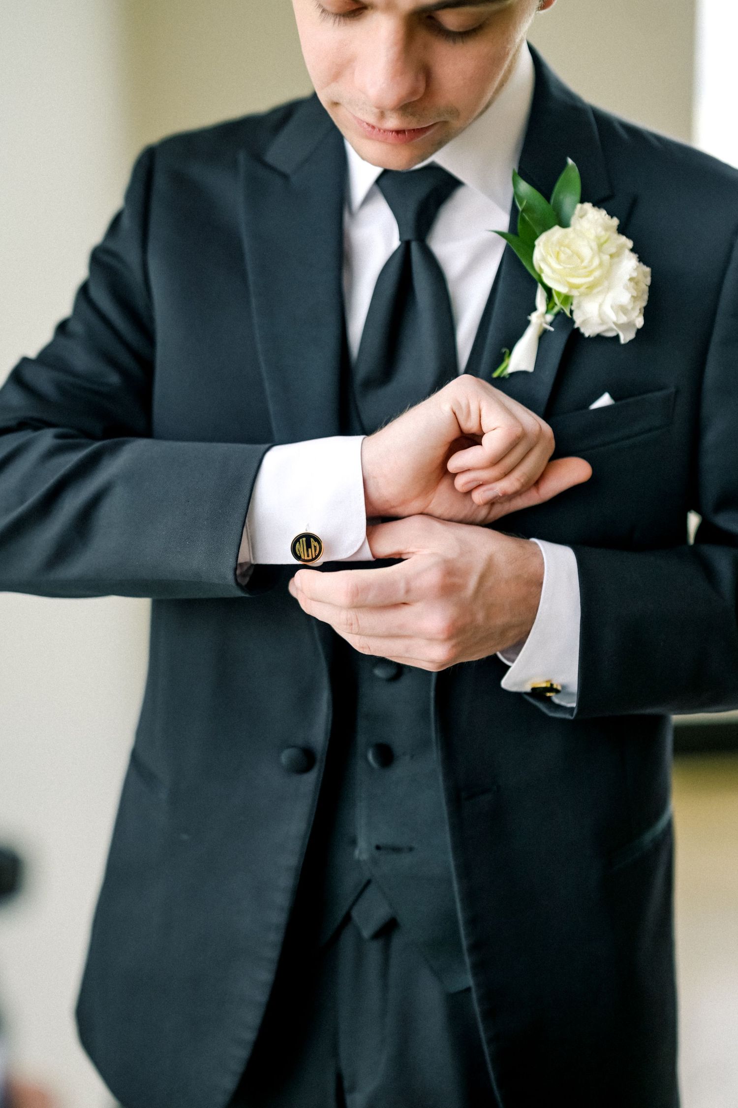 Groom in a black suit adjusting his cufflinks with a white rose boutonniere on his lapel.