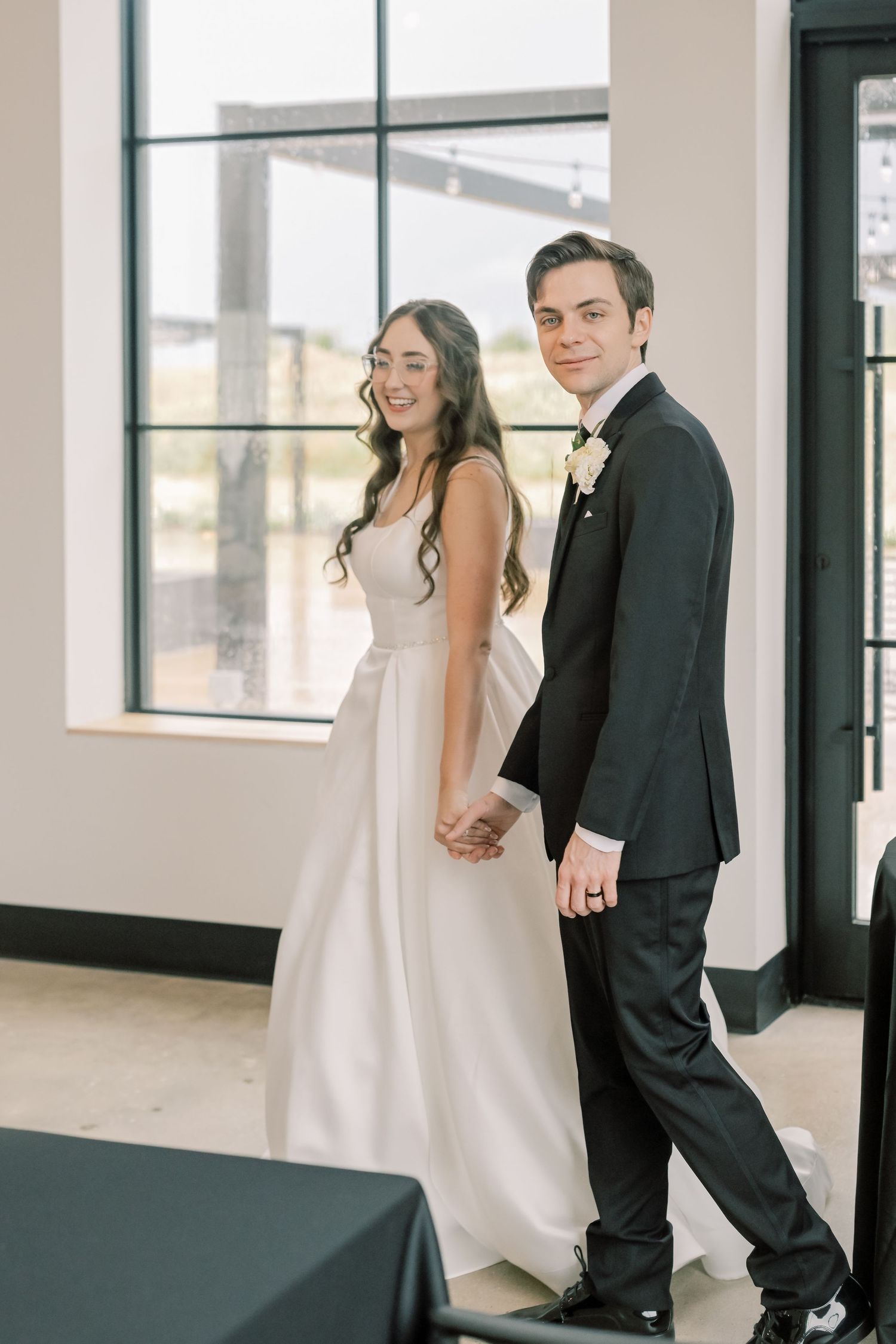 A smiling bride in a white gown and groom in a black tuxedo hold hands and pose in a bright modern venue.