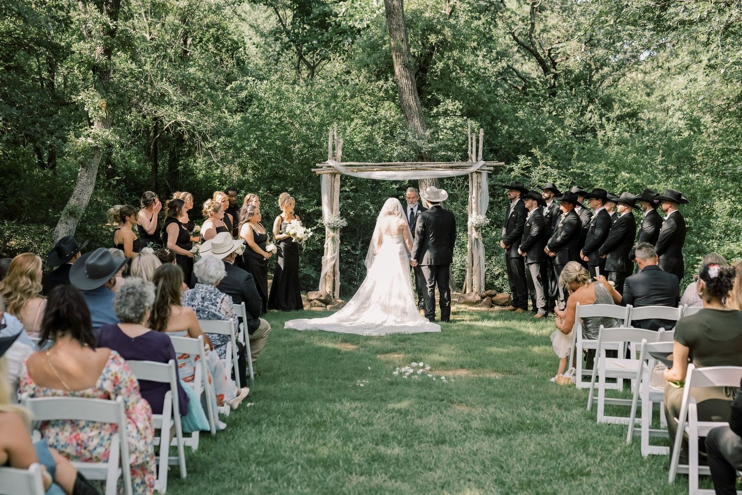 Bride and groom exchange vows at rustic wooden arch surrounded by wedding party in black attire.