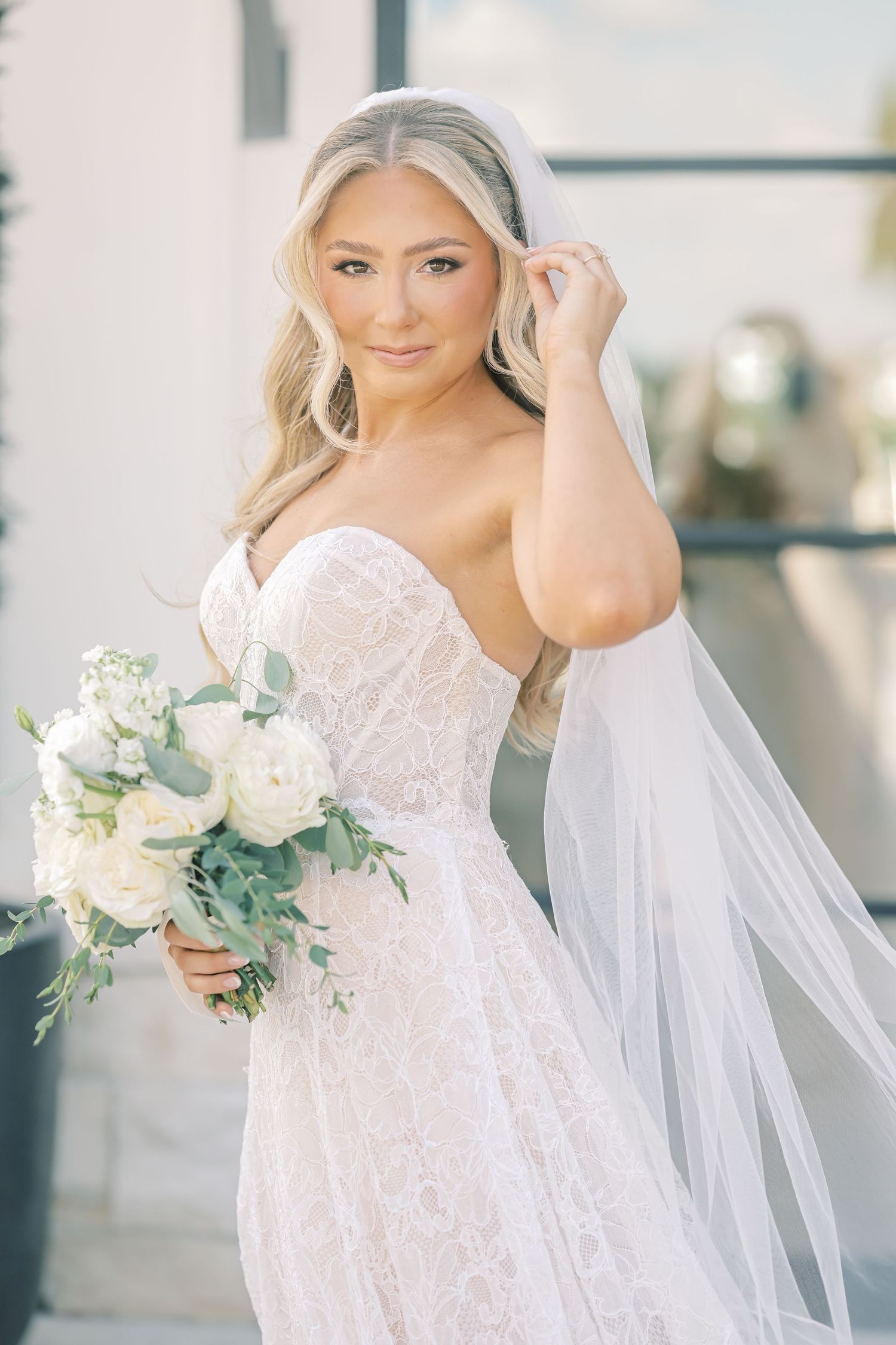 Blonde bride in strapless lace gown holding white floral bouquet with veil.