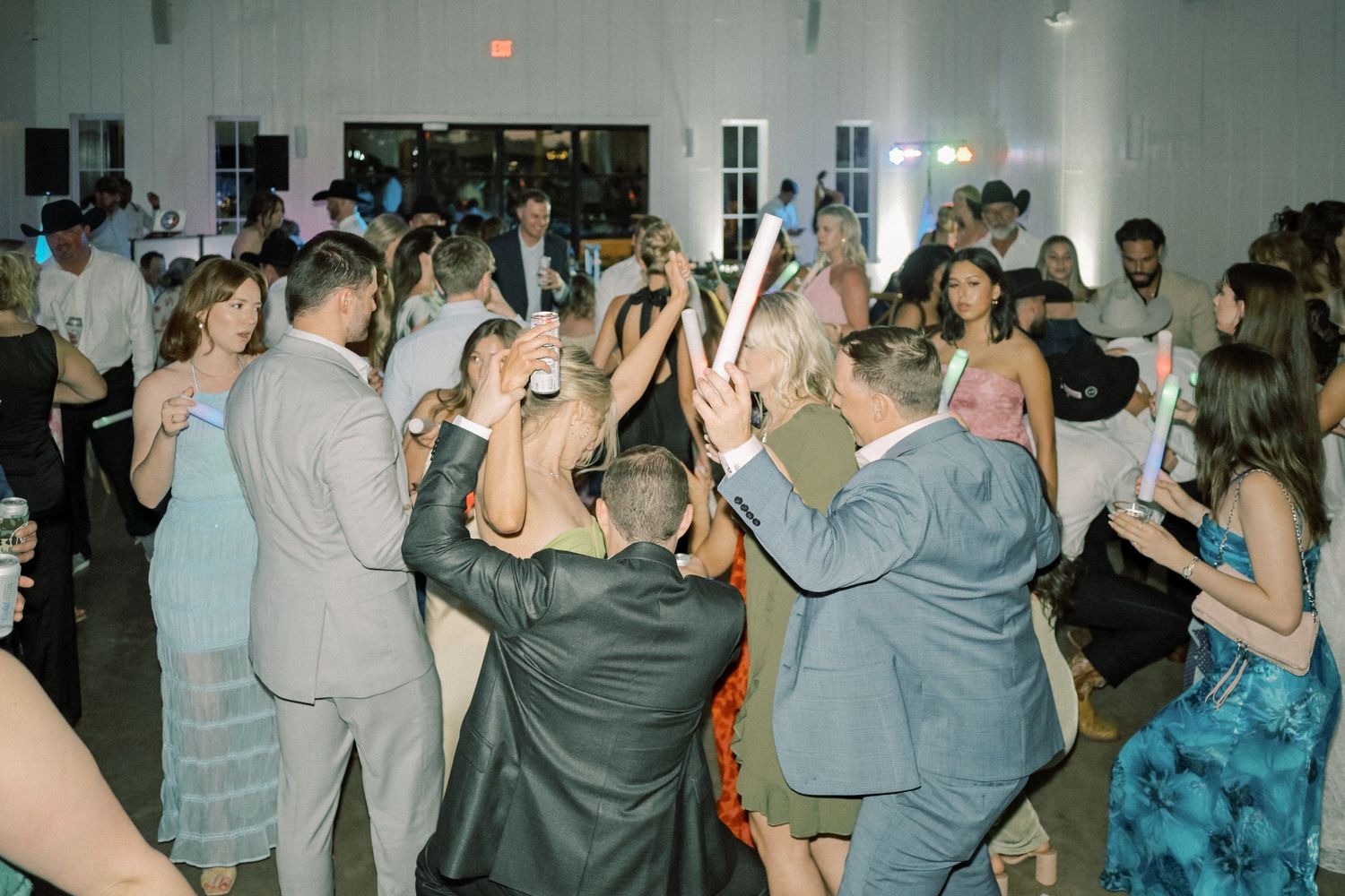 Energetic wedding reception crowd dancing and waving foam glow sticks in a bright white venue.