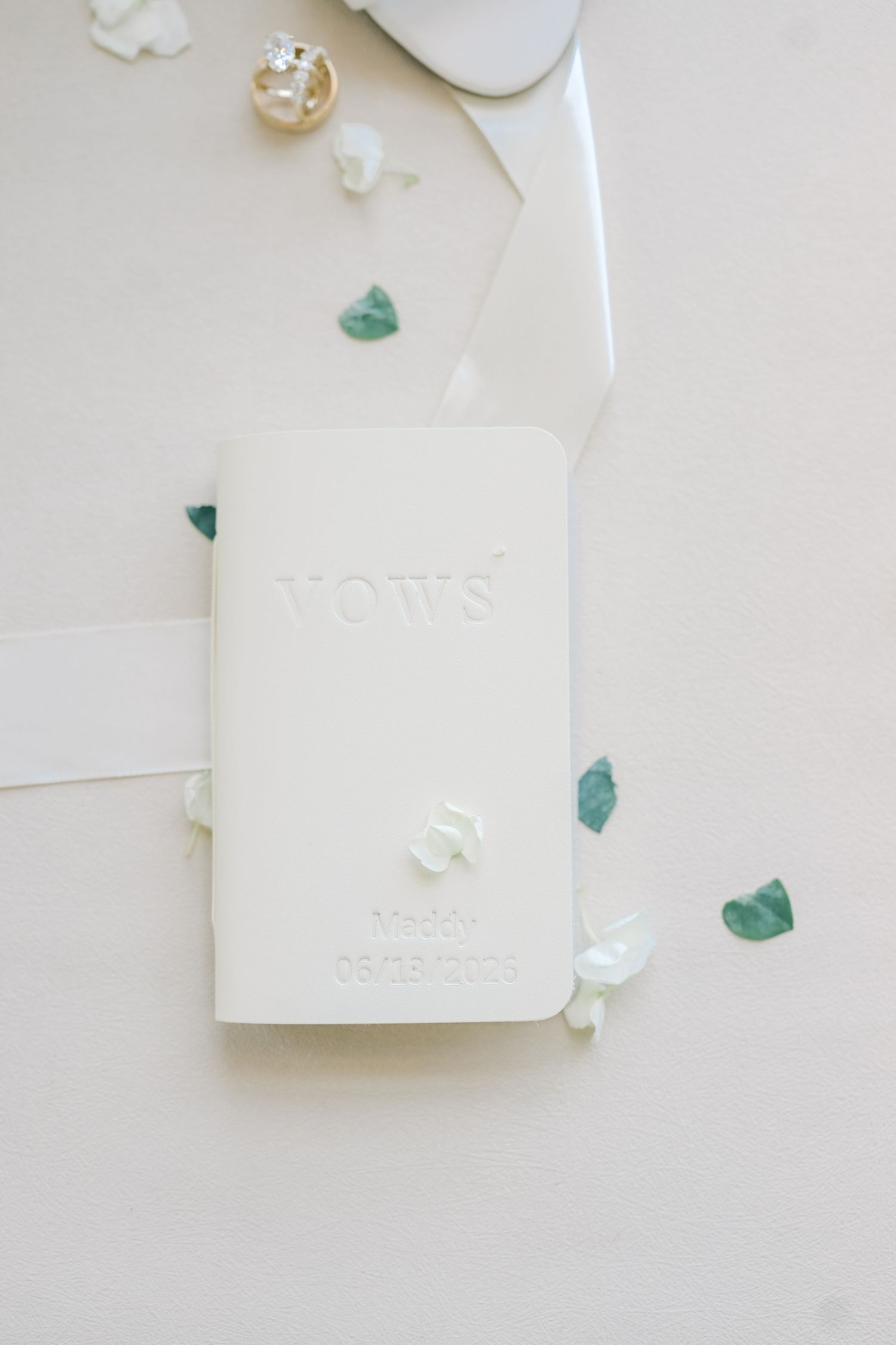 White embossed vows book with wedding date on a soft white background with green leaves and flower petals.