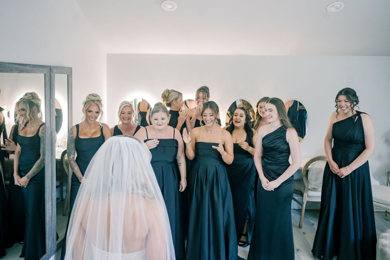 Bridesmaids in black dresses reacting with joy to bride in white veil during first look reveal.