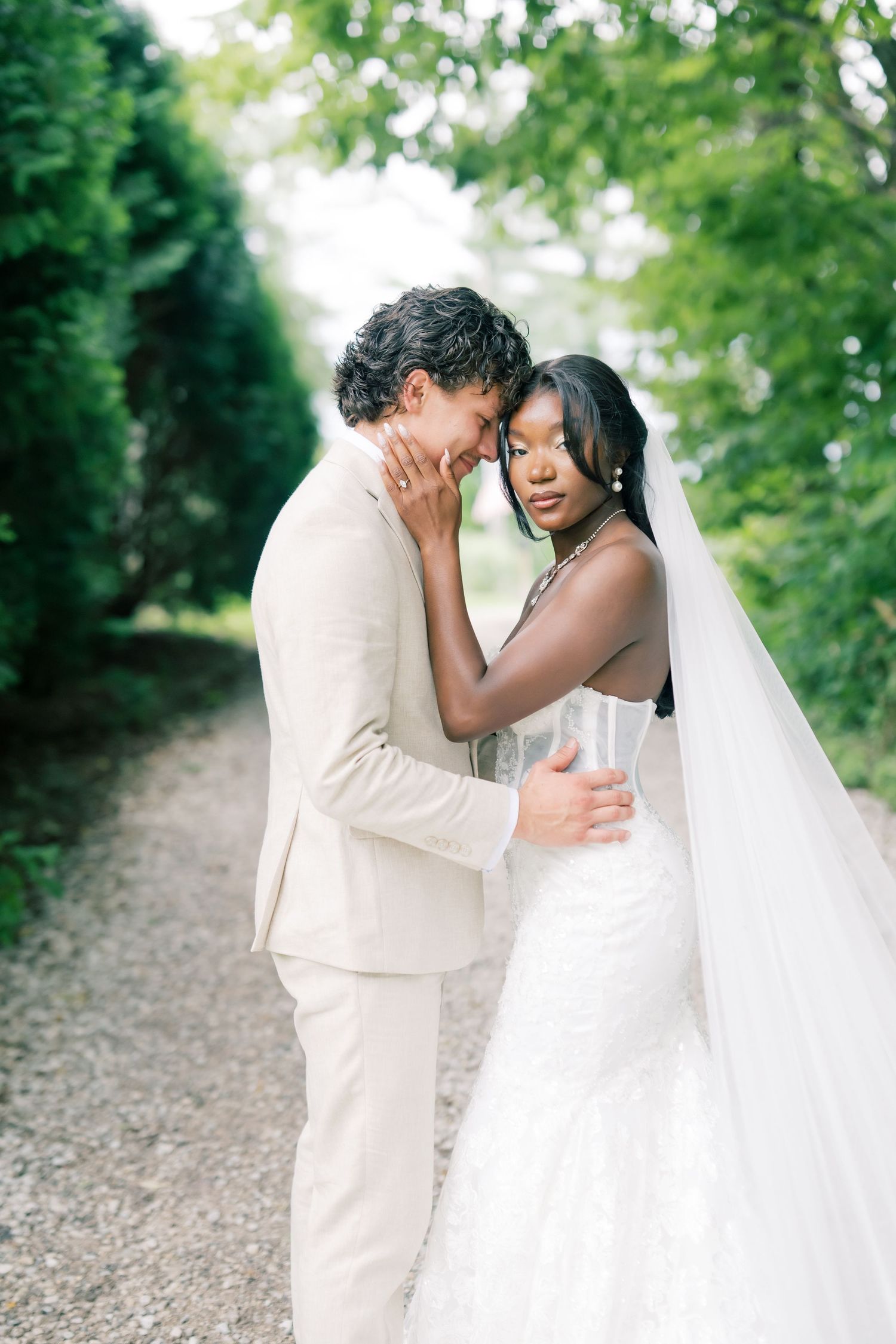 Bride and groom share an intimate forehead touch on a tree-lined gravel path, bride in lace mermaid gown.