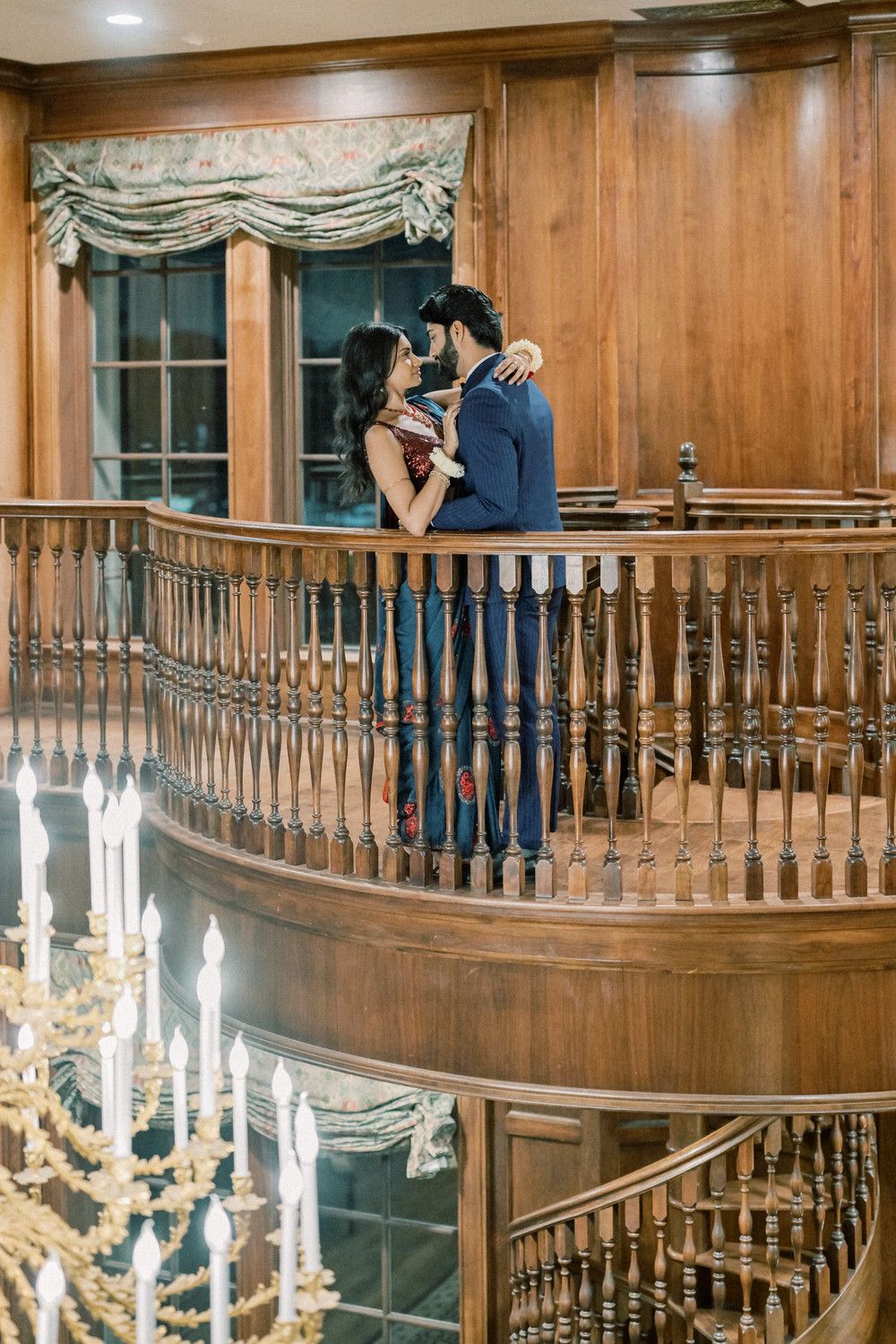 Romantic couple embracing on curved wooden balcony with chandelier in foreground.