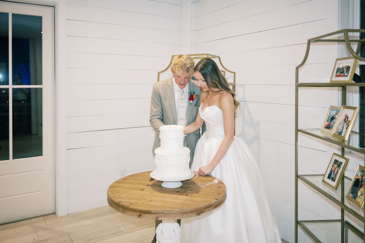 Bride and groom cutting white wedding cake on rustic wooden table in elegant white shiplap venue.