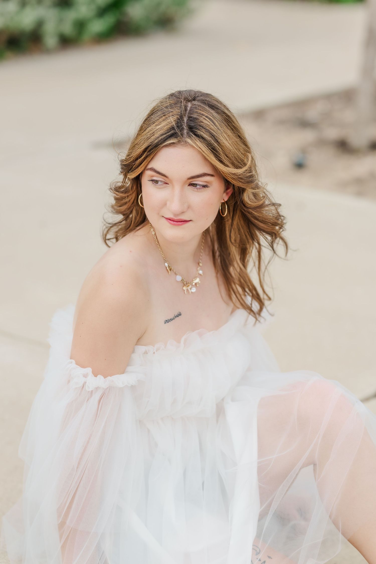 Young woman in a sheer white off-shoulder dress with a pearl necklace, sitting outdoors in soft natural light.