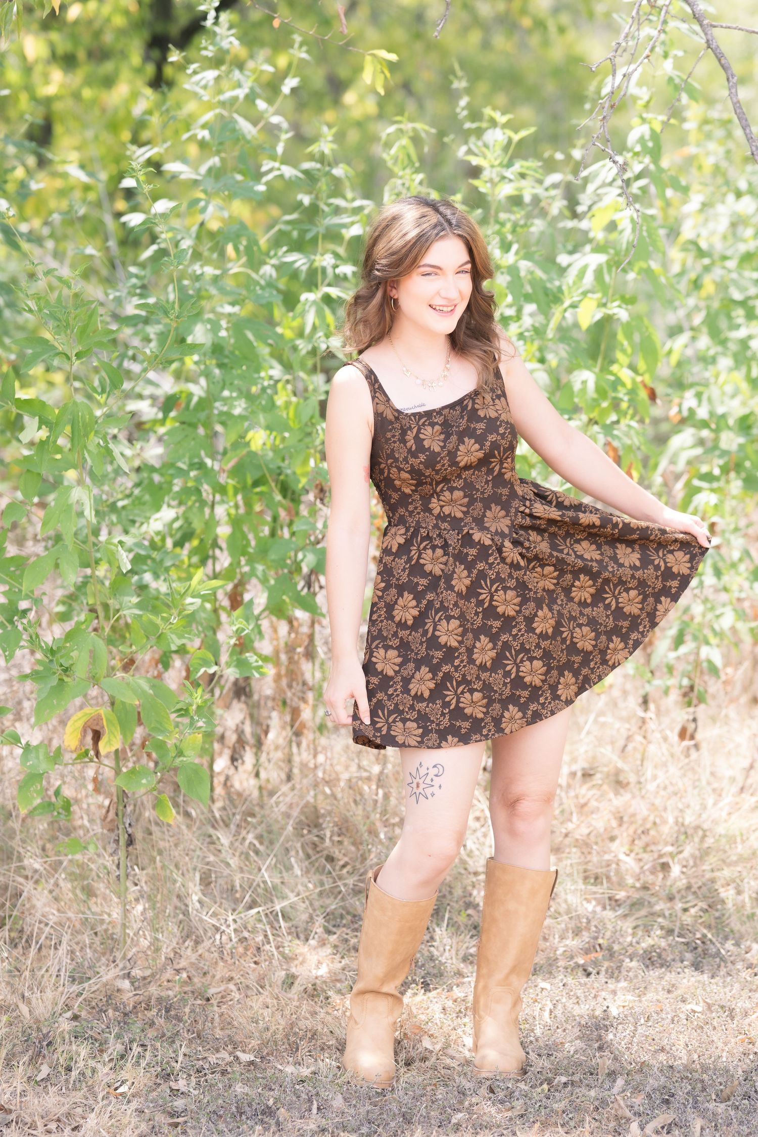 Young woman in floral dress and tan boots smiling outdoors among green trees.