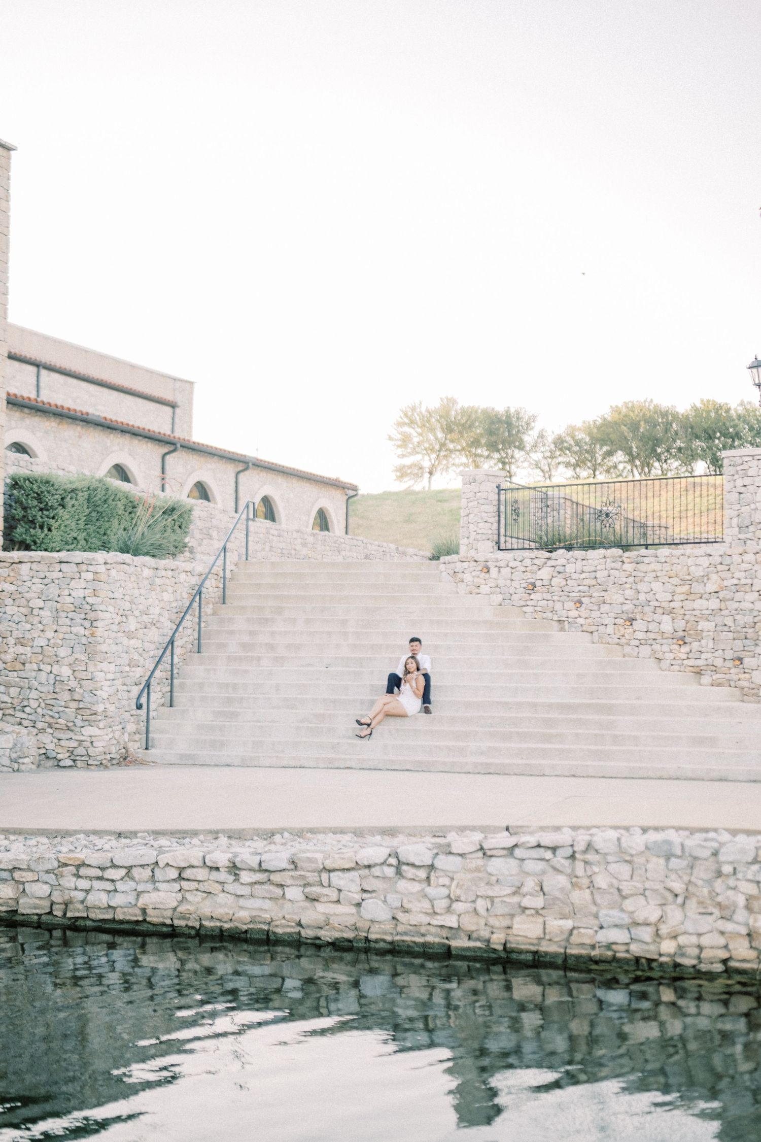 Couple sitting on stone steps by a reflective pool near a rustic building during golden hour.