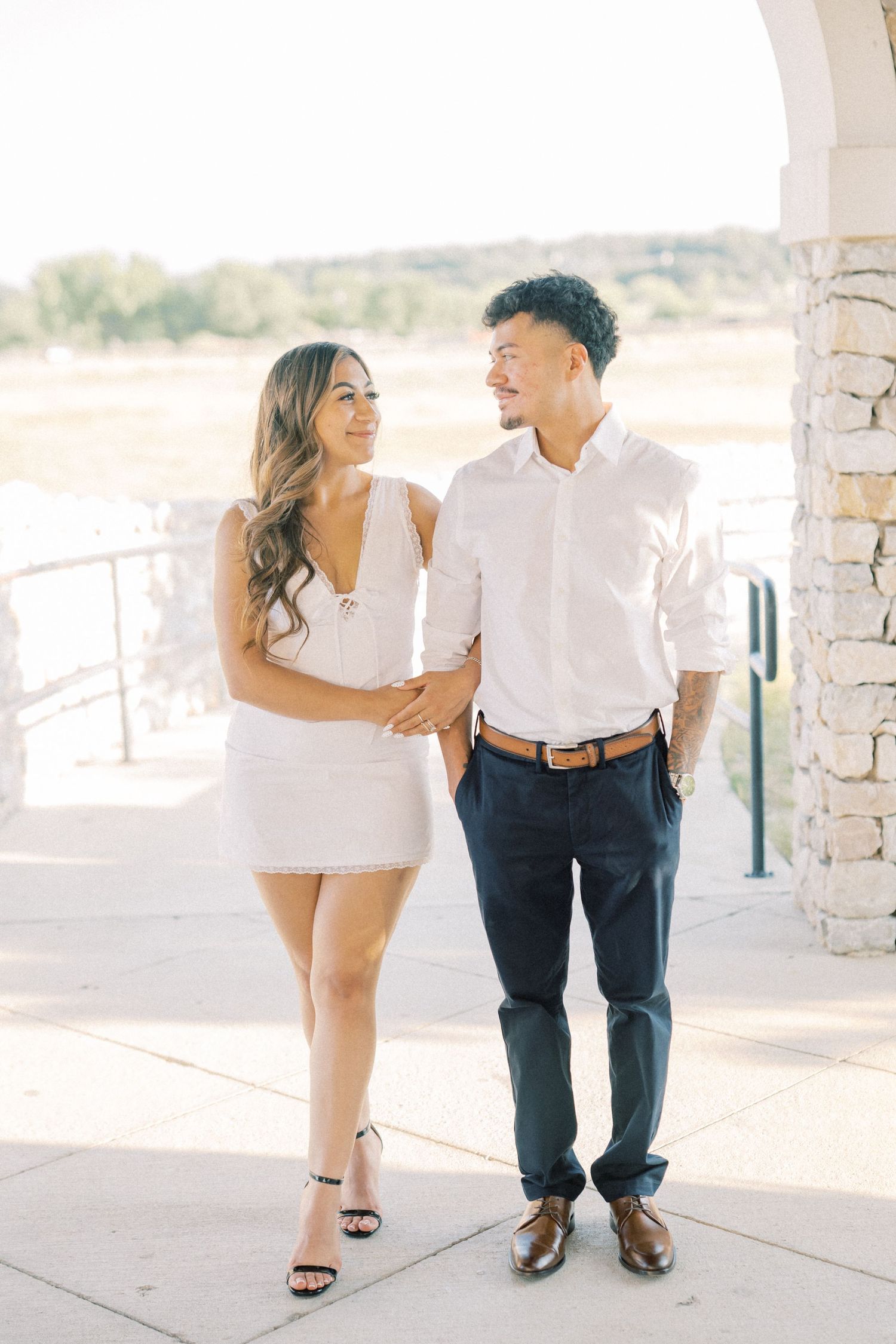 Couple in white outfits smiling at each other outdoors near stone columns and scenic waterfront.