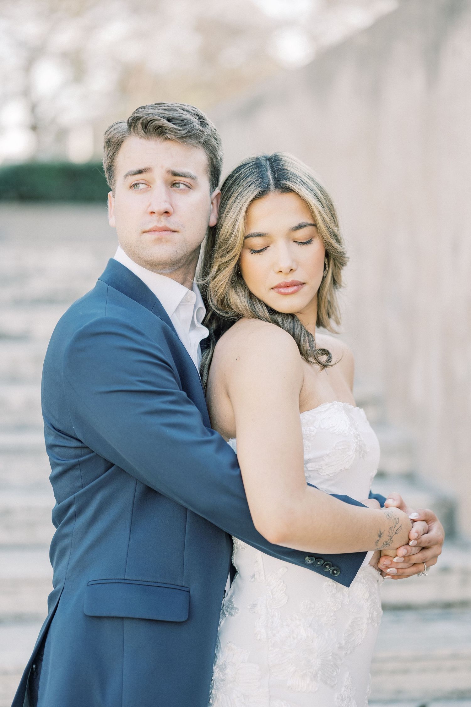 A romantic wedding portrait shows a couple embracing outdoors, with the bride in white lace and groom in navy blue suit.