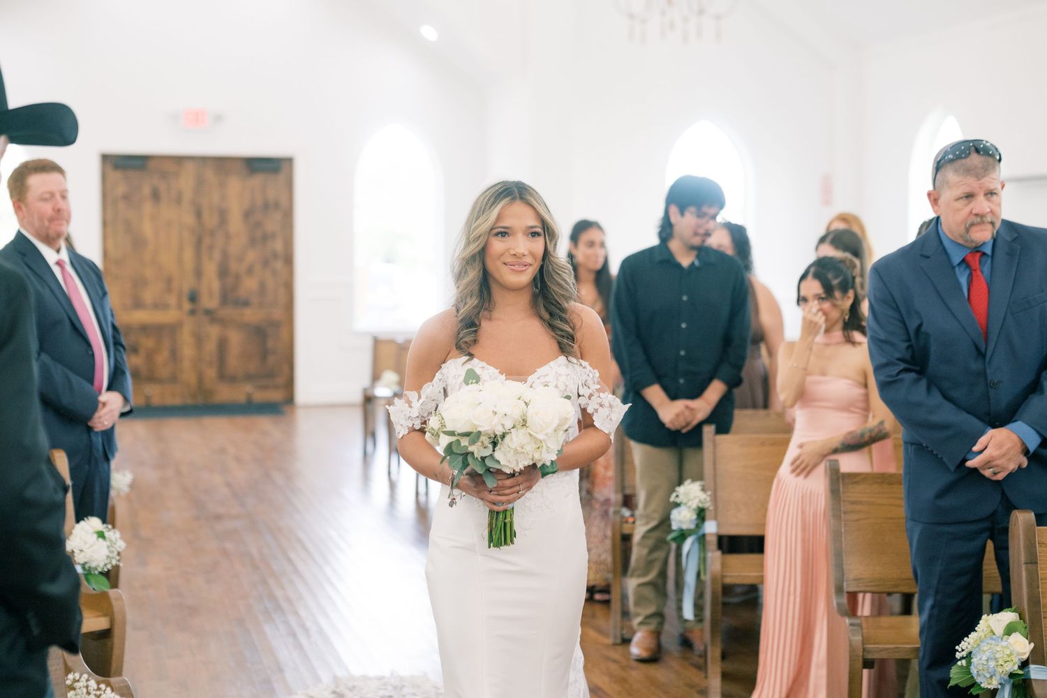 Bride walking down the aisle in a fitted lace gown holding a white bouquet in a white chapel.