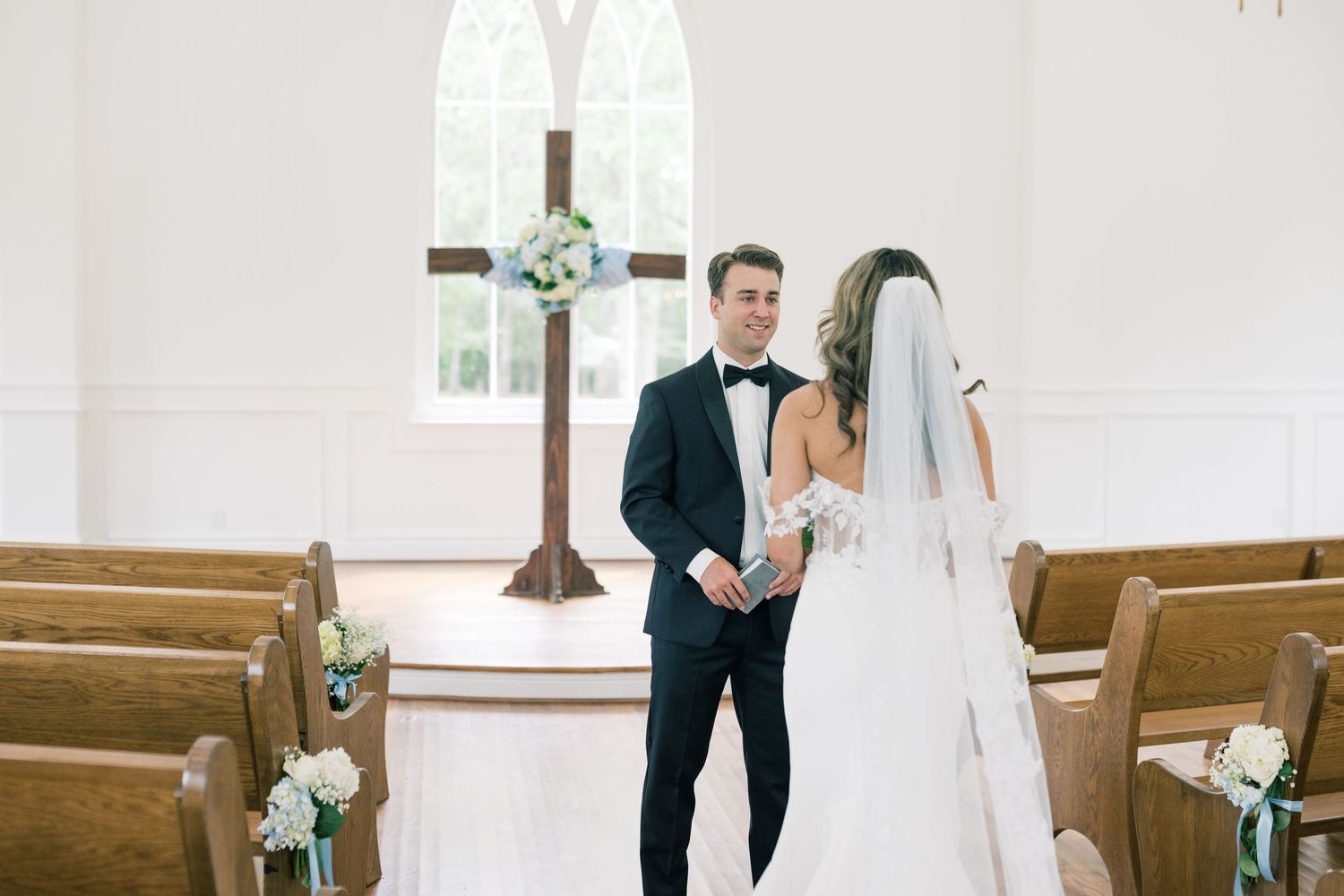 Groom in black tux awaits bride walking down chapel aisle.