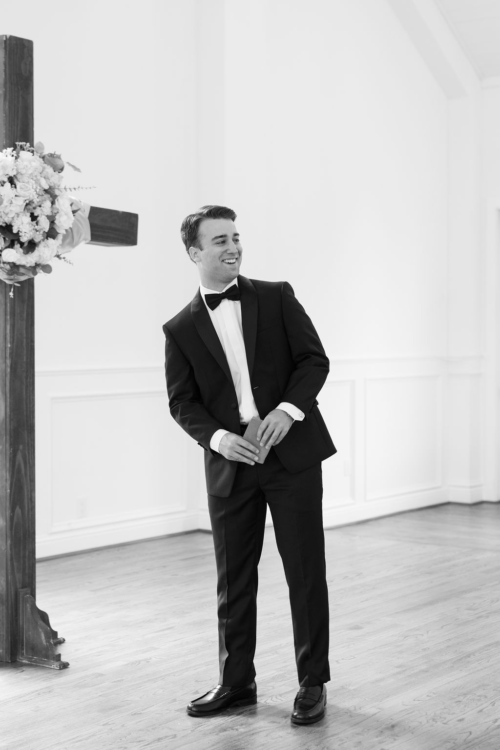 Smiling groom in black tuxedo and bow tie standing at wedding altar beside wooden cross and floral arrangement.