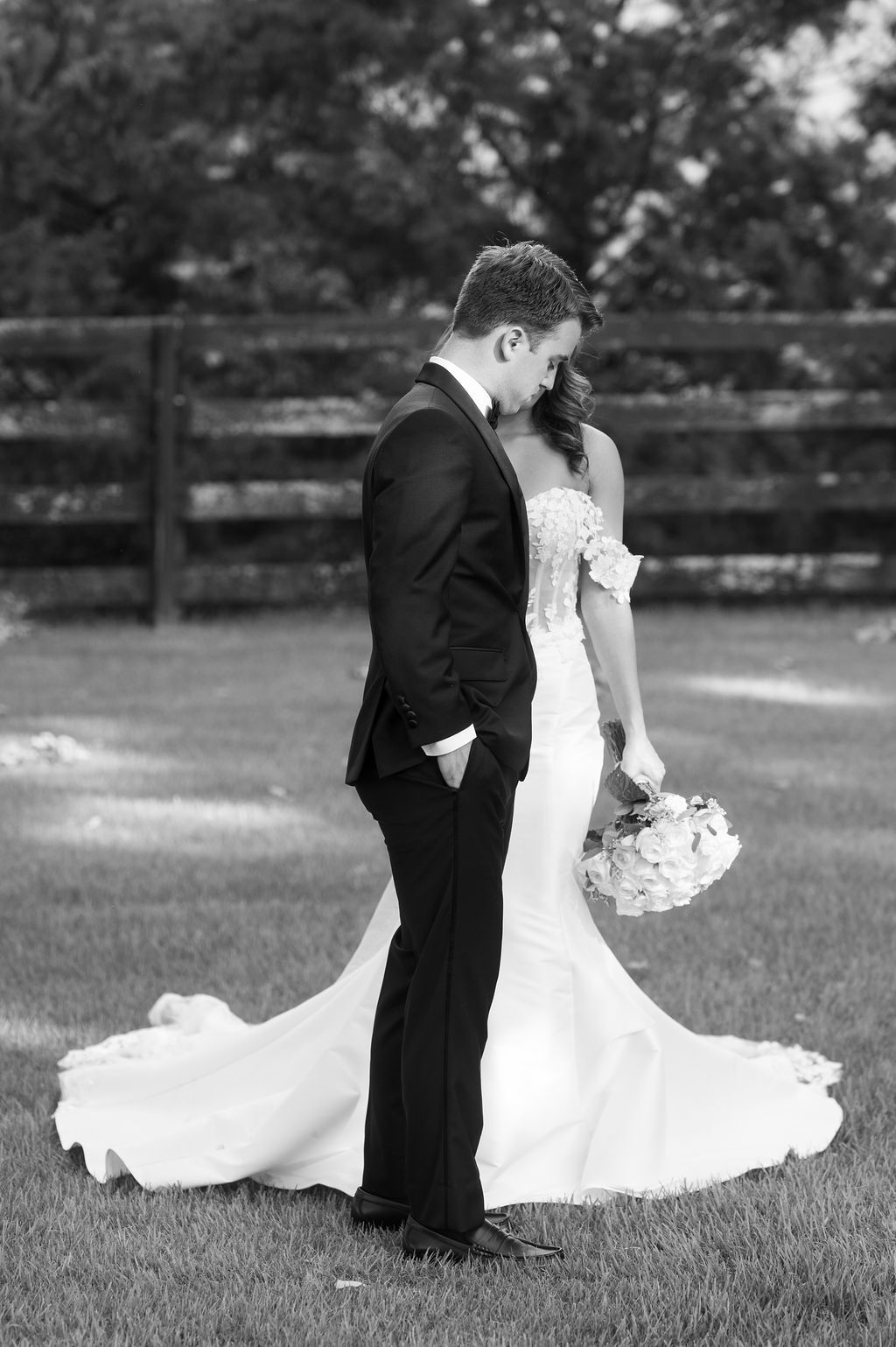 Black and white photo of bride in mermaid gown holding bouquet standing behind groom in tuxedo on lawn.