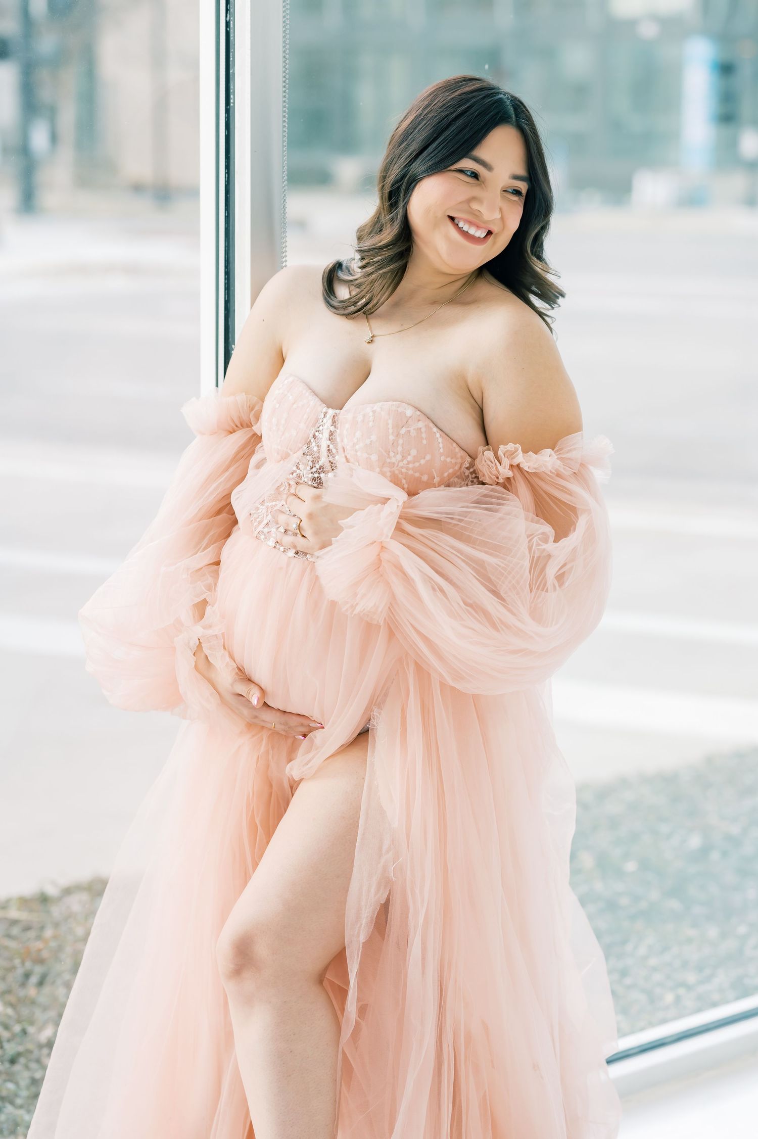 Smiling pregnant woman in a blush pink tulle maternity gown with lace bodice posing by a large window.